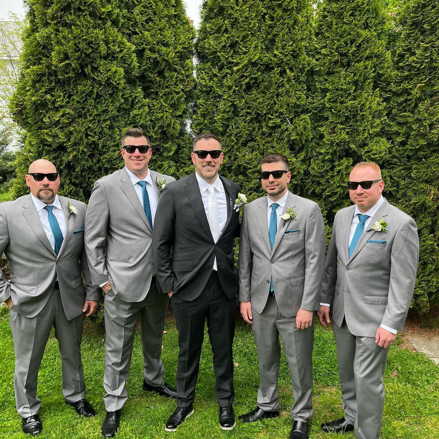 Five men wearing suits and sunglasses, posing outdoors in front of tall evergreen trees. Four are in grey suits with teal ties and one in a black suit with a white tie, all with boutonnières.