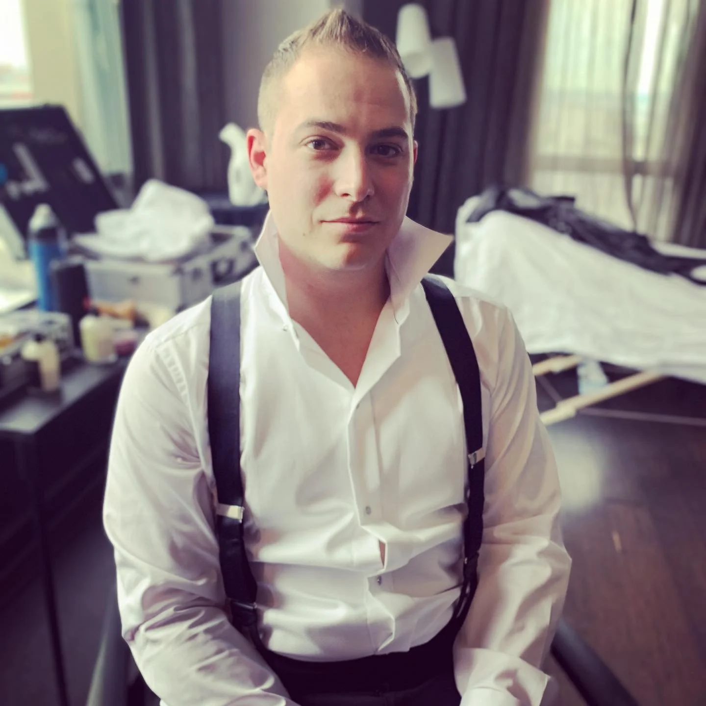 Person in white shirt with suspenders sitting in a room with various items in the background, including clothes on a rack and grooming products on a table.