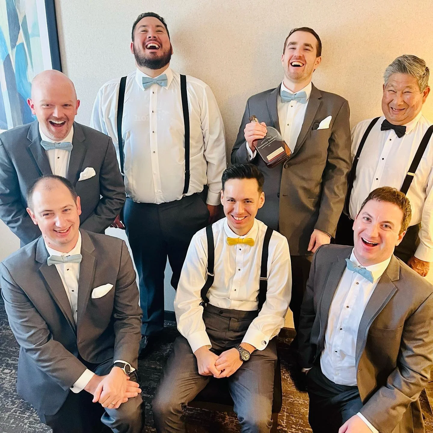 A group of seven men in suits and bow ties, laughing and posing indoors. One is holding a liquor bottle, and some are wearing suspenders. They appear to be at a wedding or formal event.