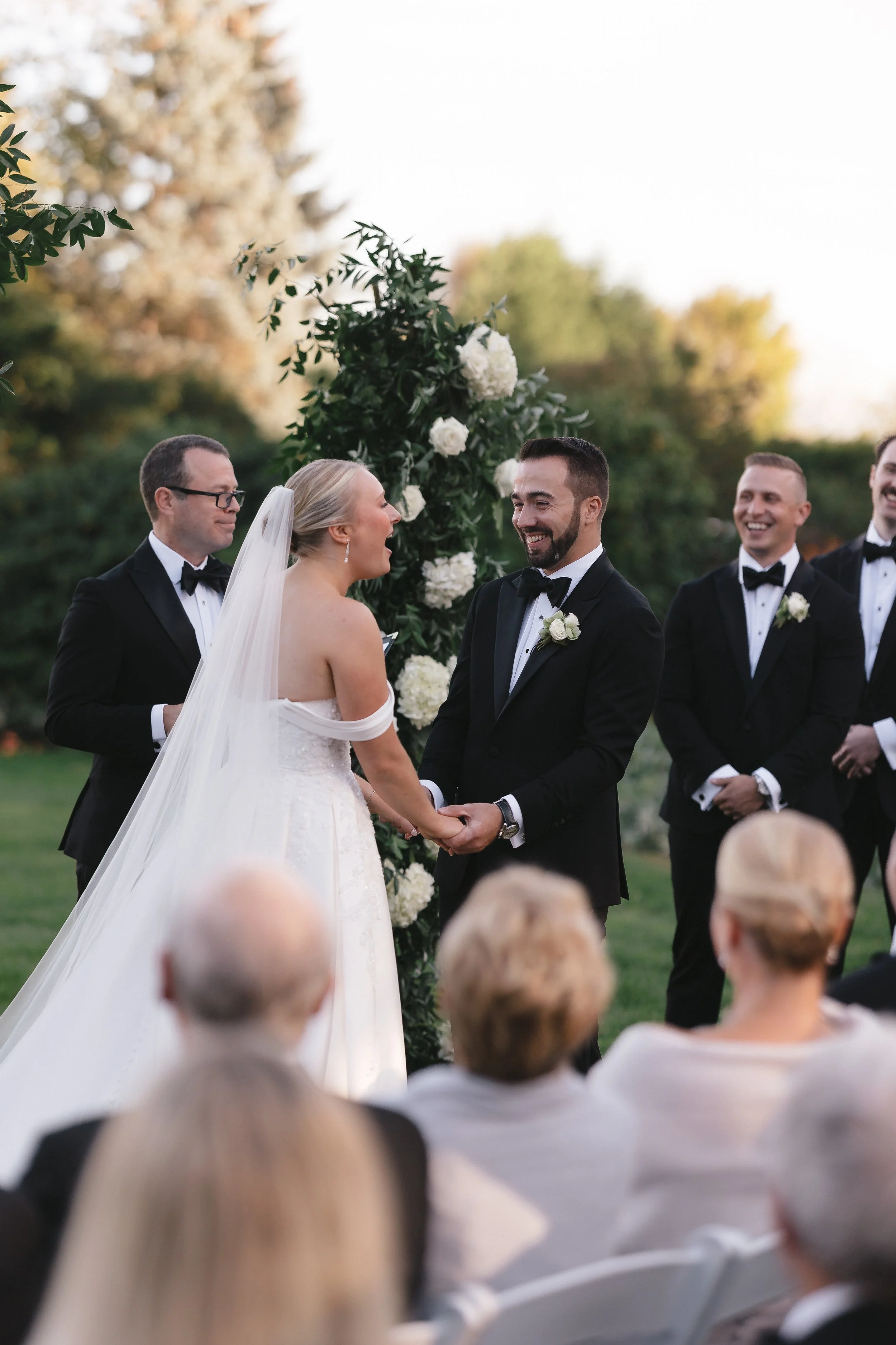 A wedding ceremony outdoors with a bride and groom holding hands and smiling, surrounded by groomsmen and guests, with a floral arch and trees in the background.