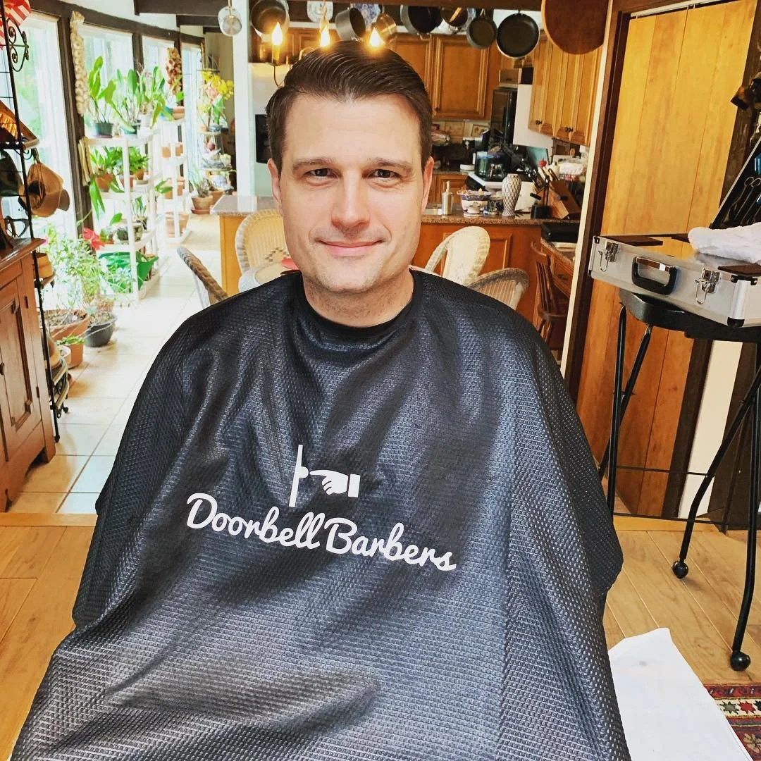 A person wearing a black salon cape with the words "Doorbell Barbers" on it, sitting in a kitchen with plants and cooking utensils visible in the background.