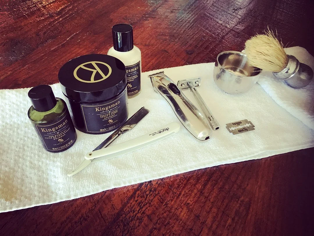 Shaving kit with Kingsman grooming products, razor, blade, brush, and towel on wooden surface.