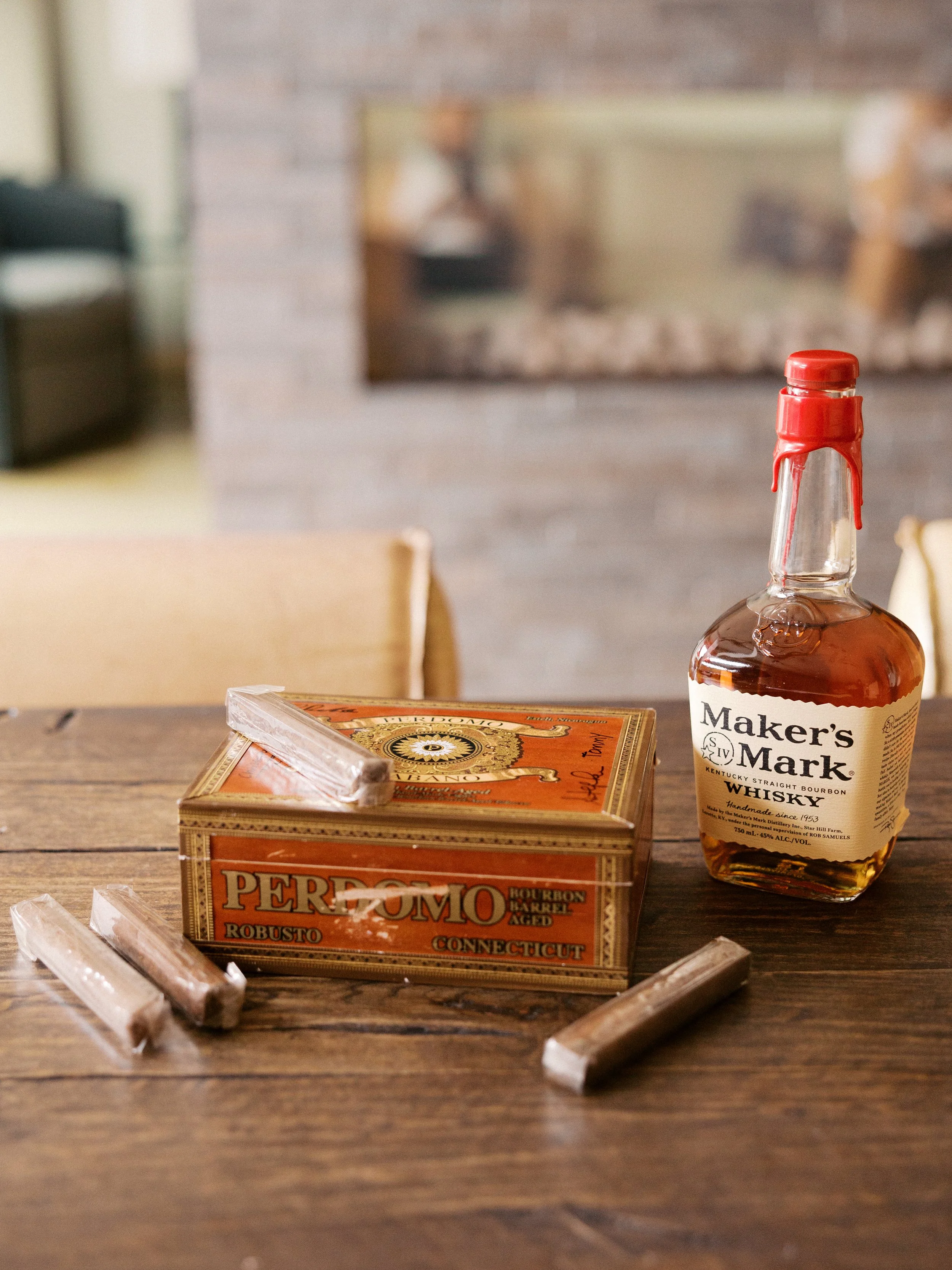 Bottle of Maker's Mark whiskey beside a Perdomo cigar box and cigars on a wooden table, with a blurred background.