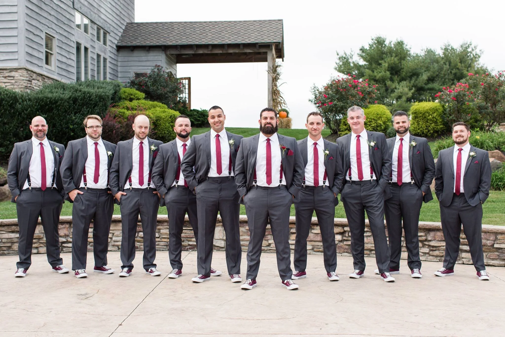 A group of men in matching gray suits and red ties, standing outdoors in front of a stone and wooden building. They are wearing sneakers and have boutonnieres on their lapels, indicating they might be groomsmen at a wedding. The background features g