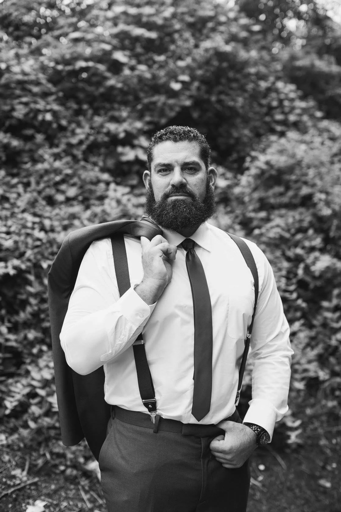 Man with beard in formal attire, holding jacket over shoulder, outdoors in black and white.