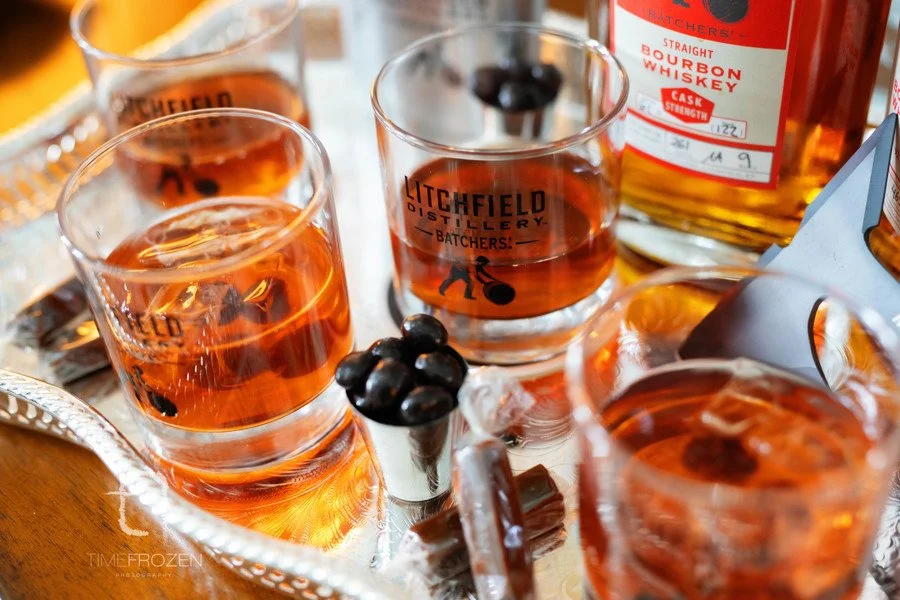 Glasses of amber-colored whiskey on a tray with black candies, featuring Litchfield Distillery branding and a bottle of straight bourbon whiskey in the background.