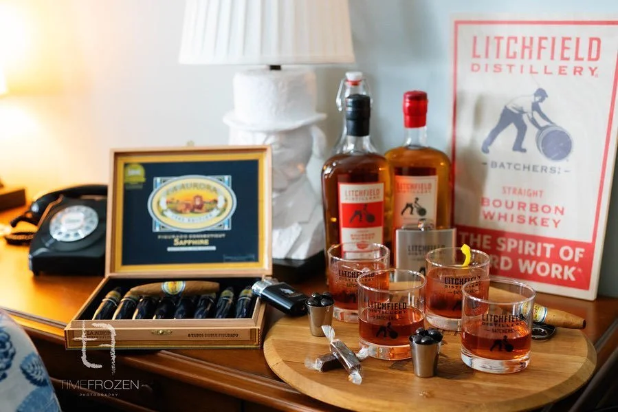 A setup featuring two bottles of Litchfield Distillery bourbon, a box of cigars open on a table, and a wooden platter with four glasses of whiskey and two small jiggers. A vintage rotary phone and a lamp with a hat-shaped base are in the background, 