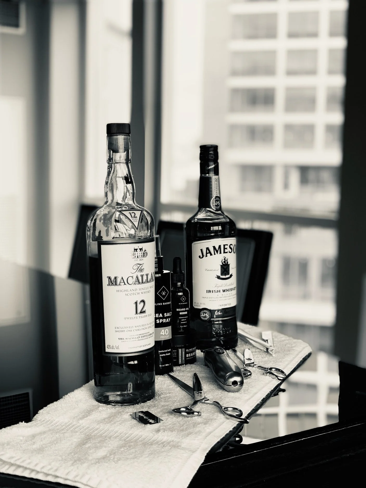 Two whiskey bottles, grooming tools including scissors, a trimmer, and grooming products on a towel-covered surface in a room with a window view of a building.