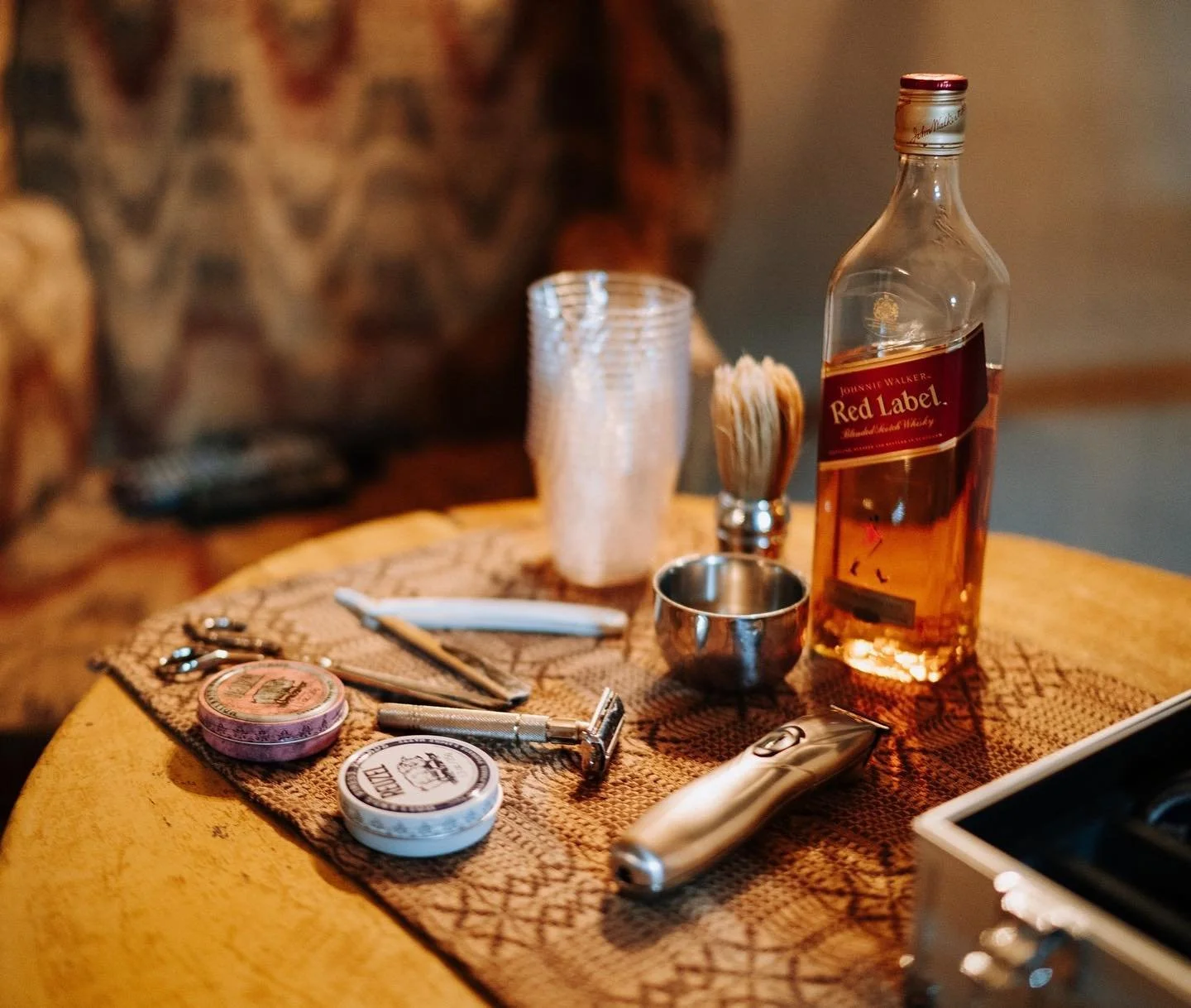 A bottle of Johnnie Walker Red Label whiskey on a table with grooming tools, including a razor, shaving cream tins, a comb, a trimmer, a shaving brush, and cups.