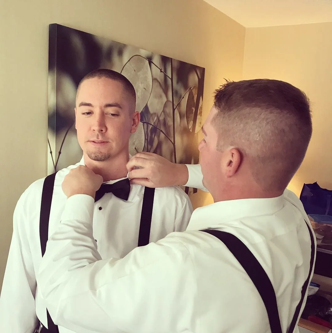 Two men in formal attire, one adjusting the other's bow tie, preparing for an event in a room with artwork on the wall.
