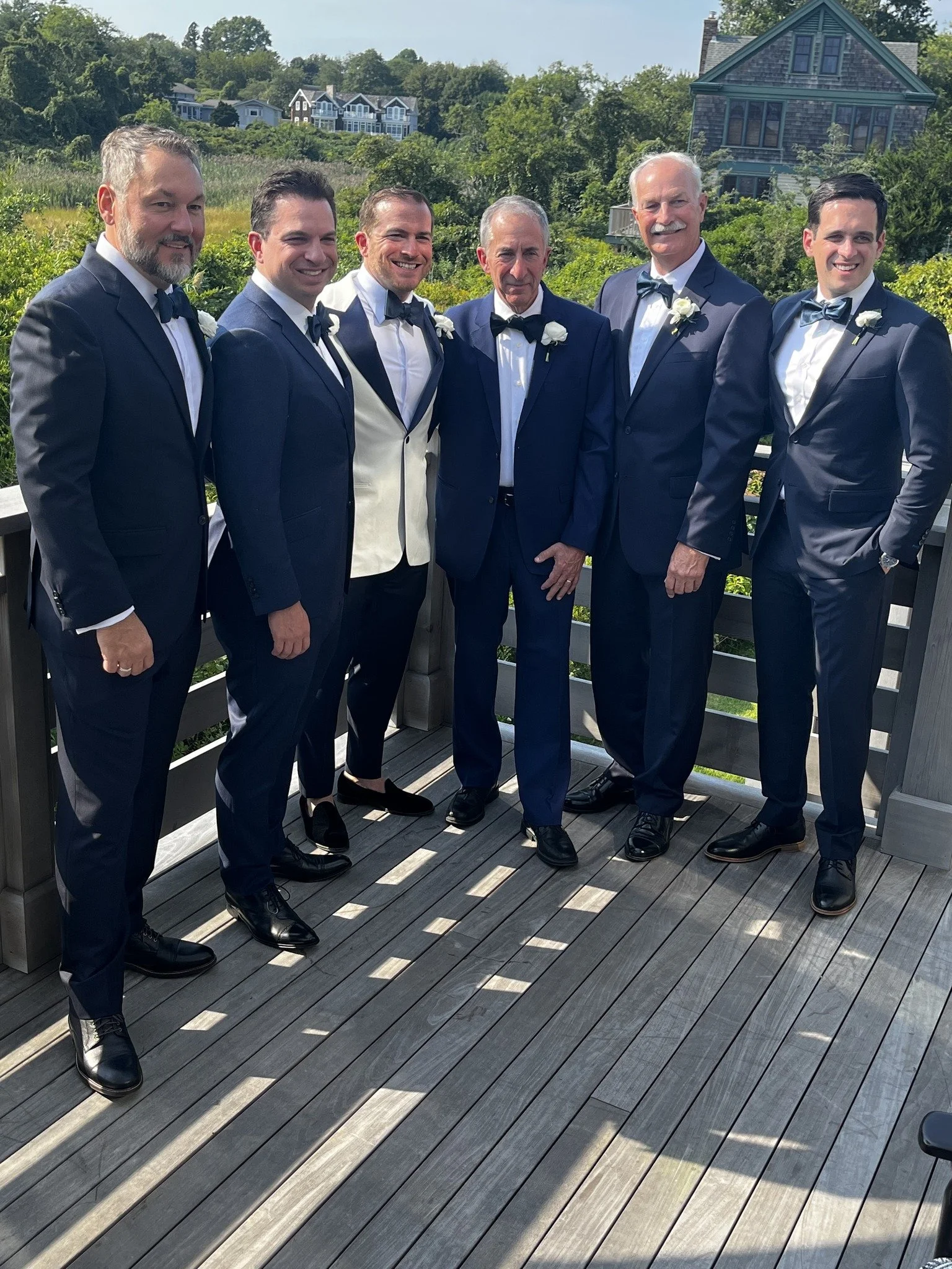 Six men in formal suits standing on a wooden deck, posing for a photo with greenery and houses in the background, sunny day.