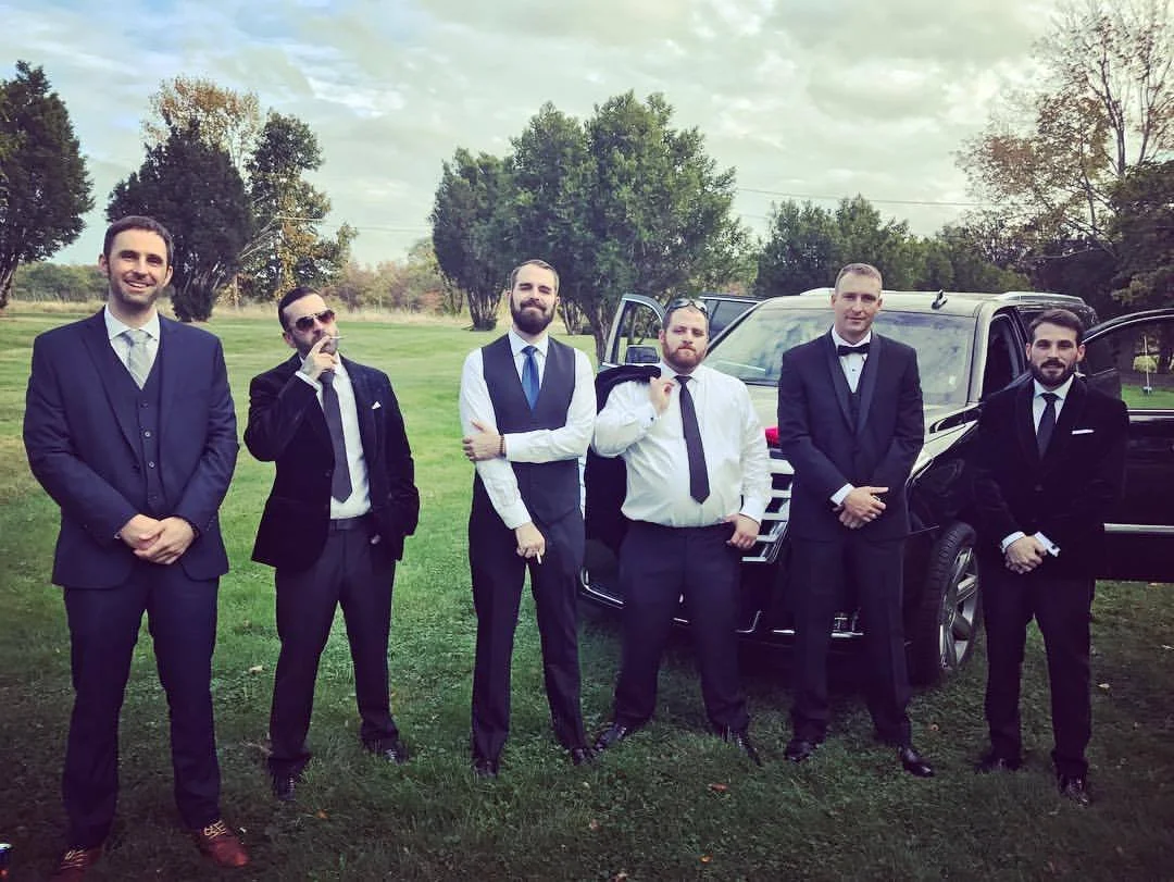 Group of six men in formal attire standing in front of a black SUV on a grassy area with trees in the background.