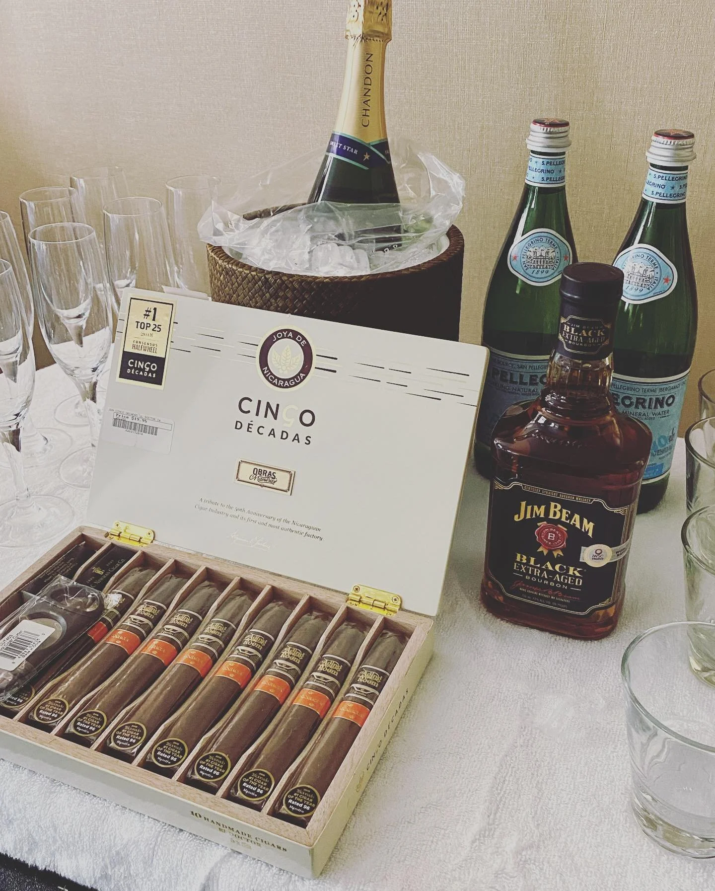 Table setting with a box of cigars labeled 'Cinco Décadas,' a bottle of Jim Beam Black Extra-Aged bourbon, a chilled Chandon champagne bottle, San Pellegrino mineral water bottles, and various glassware on a white tablecloth.