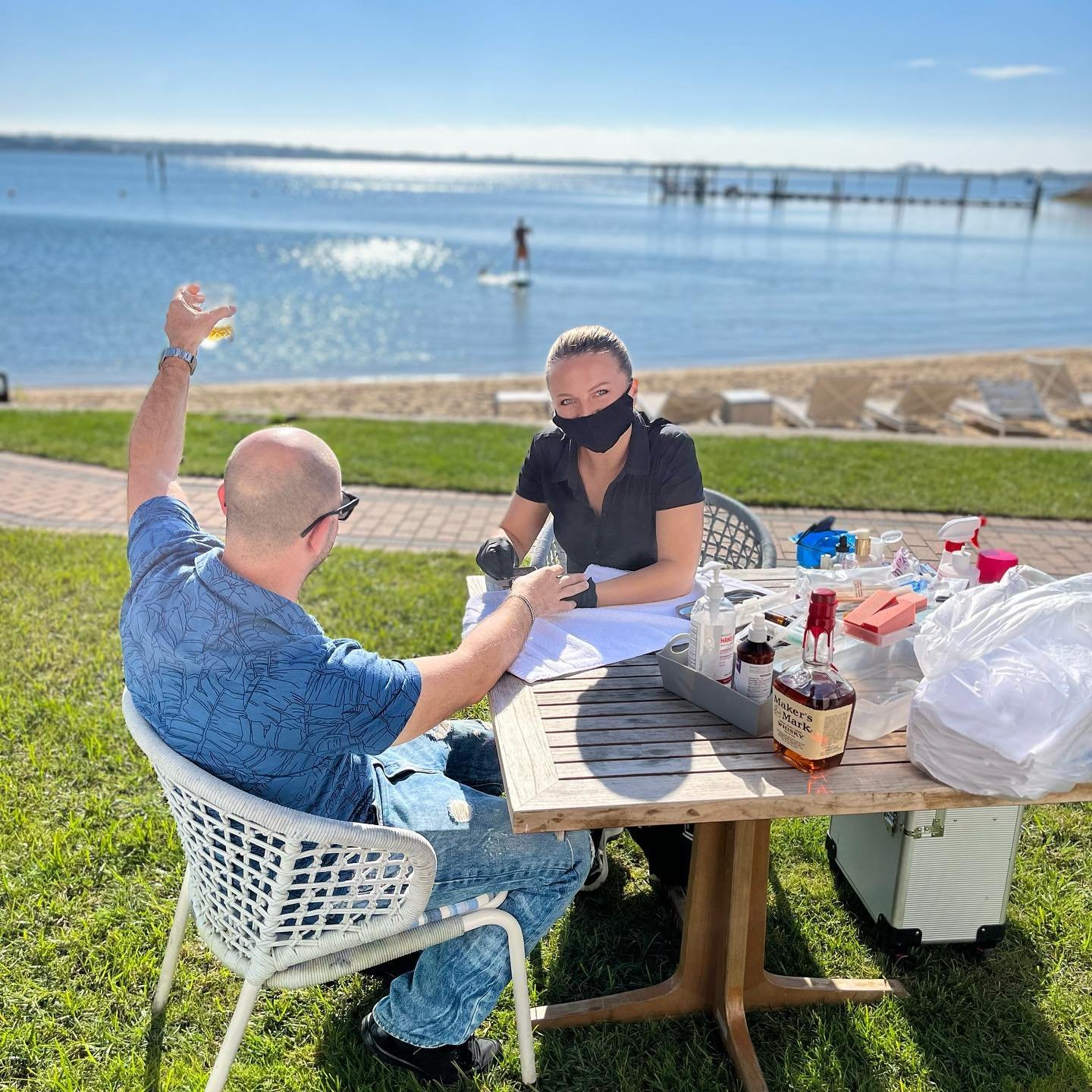 Man getting a manicure outdoors by a woman in a mask, seated at a wooden table near a beach with a paddleboarder in the background.