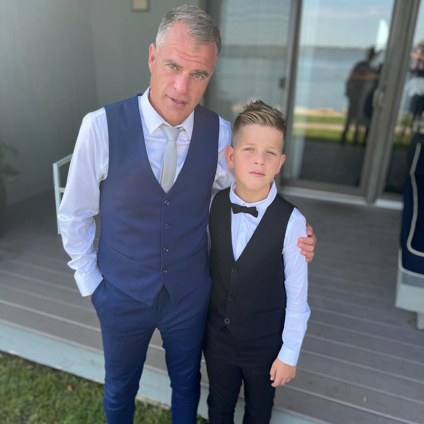 Man and child wearing formal vests and dress shirts standing on a porch with water view.