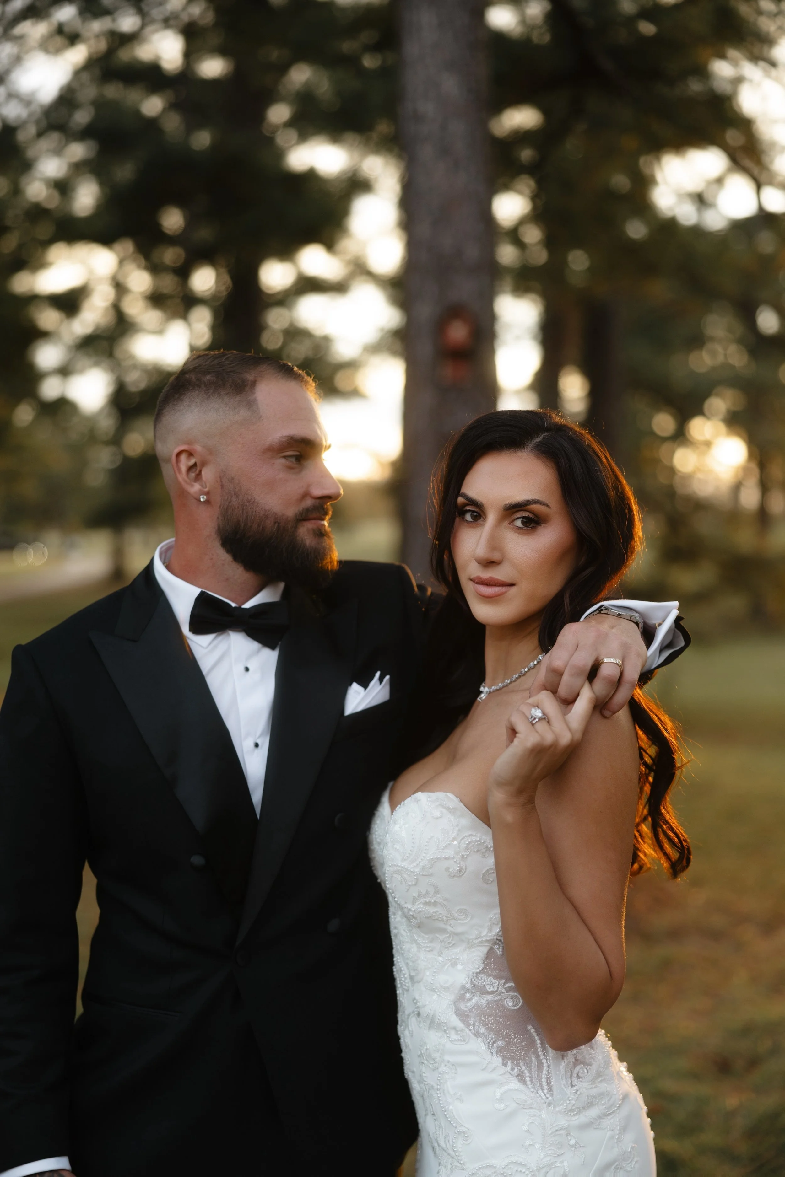 A newlywed couple dressed in wedding attire standing outdoors during sunset with trees in the background. The groom is wearing a black tuxedo with a bow tie, and the bride is in a white lace wedding gown with jewelry.