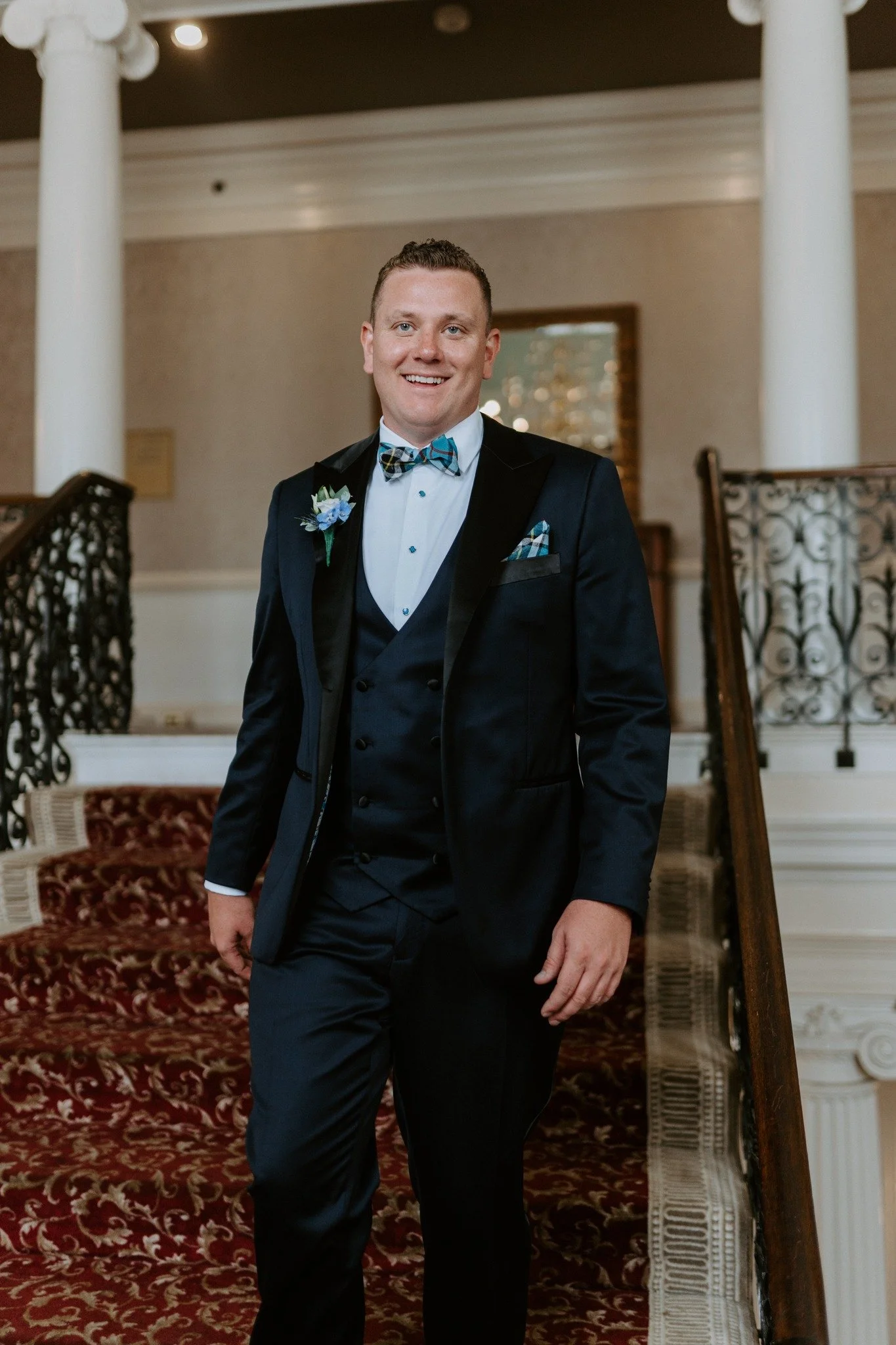 A man in a formal suit with a bow tie standing on a staircase with red carpet and ornate railings.