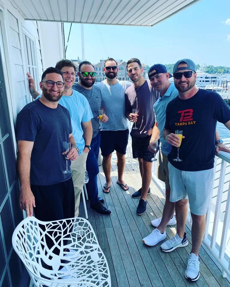 Group of seven men standing on a balcony holding drinks, with a marina and buildings in the background.