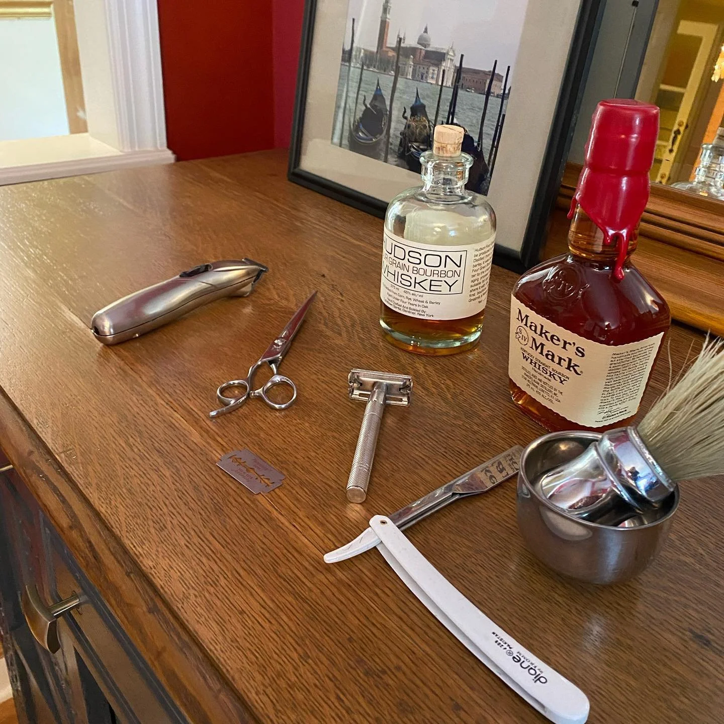 Barber tools and whiskey bottles on a wooden table, including clippers, scissors, safety razor, straight razor, shaving brush, and a framed picture in the background.
