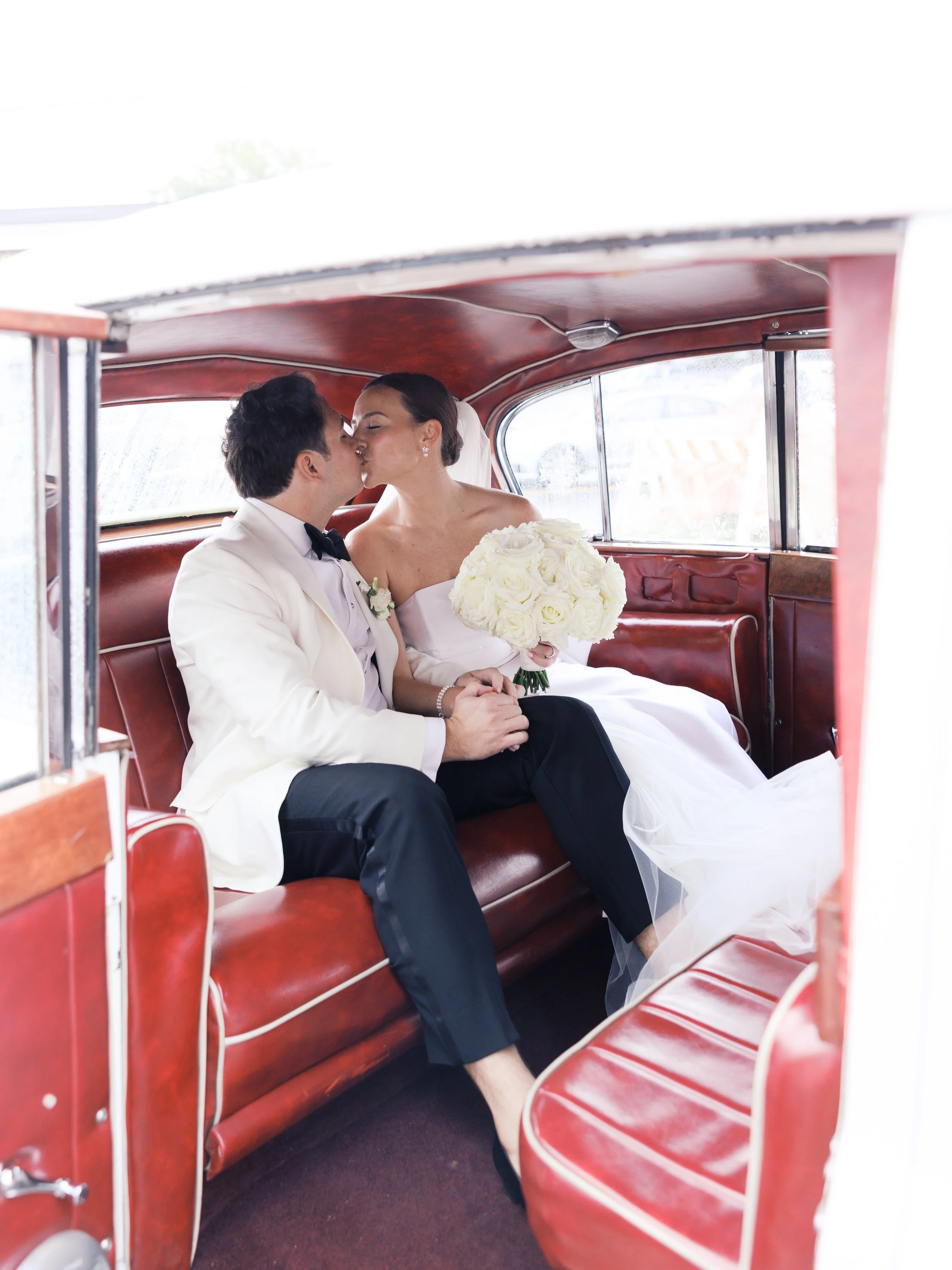 Bride and groom kissing in the backseat of a vintage car with red interior. The bride holds a bouquet of white roses.