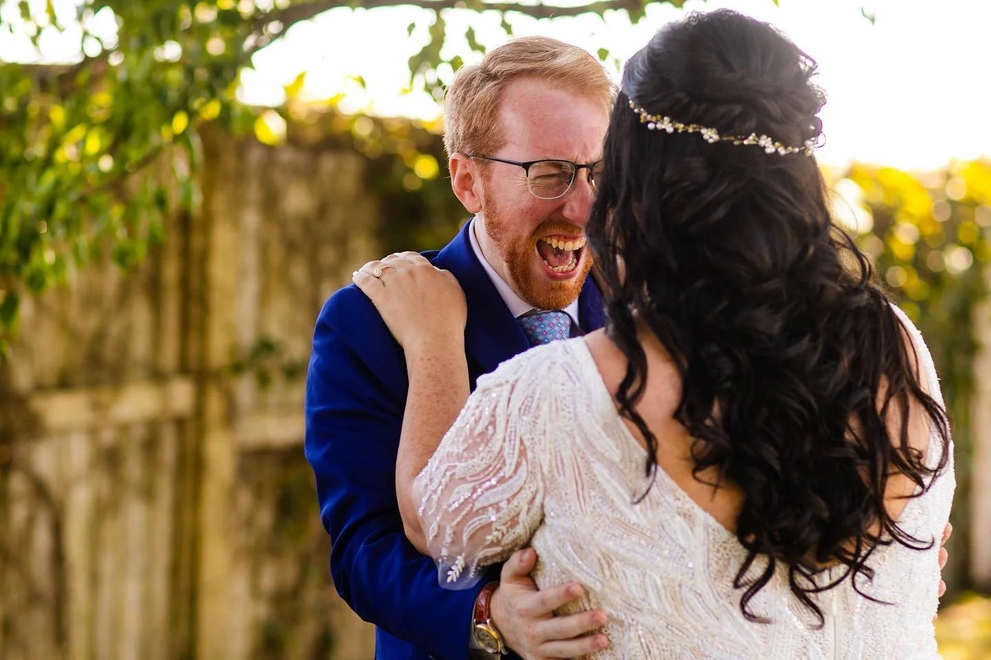 A joyful groom in a blue suit laughing while holding his bride, who has long dark hair and is wearing a beaded white dress, in an outdoor setting with greenery.