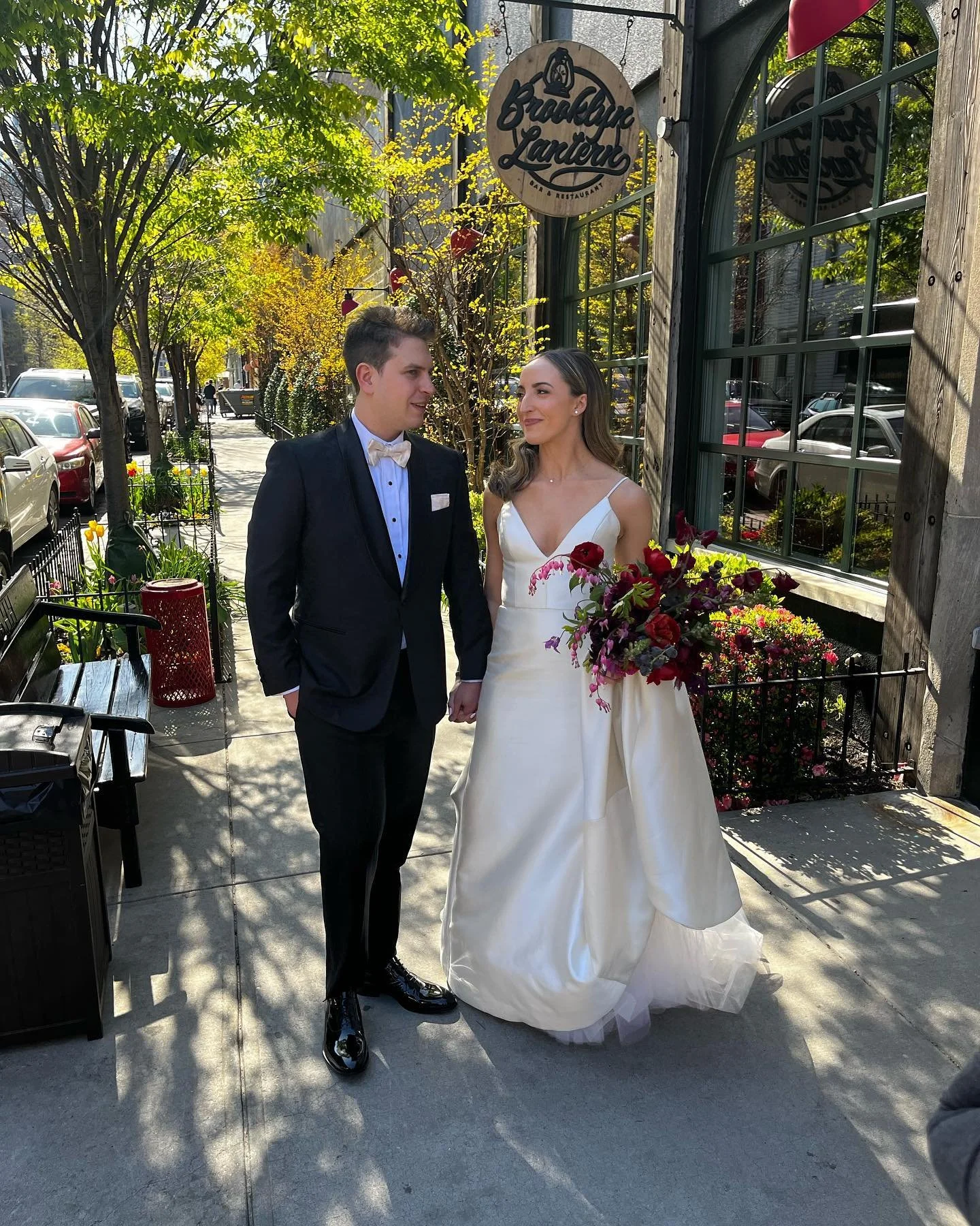 Bride and groom on a sidewalk outside a restaurant, the bride in a white dress holding a bouquet of red and pink flowers, the groom in a black suit with a bow tie.