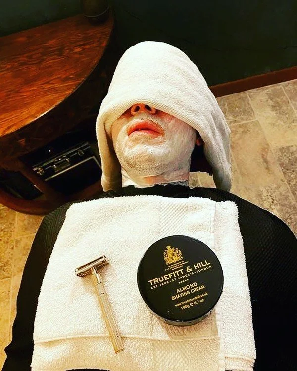 Person lying down with a towel over face and shaving cream applied, next to a razor and Truefitt & Hill almond shaving cream.
