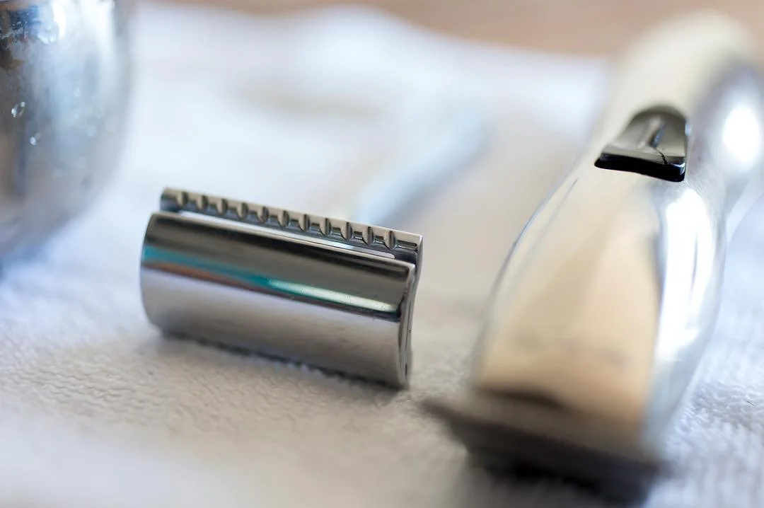 Close-up of a metal safety razor head and an electric trimmer on a white towel.