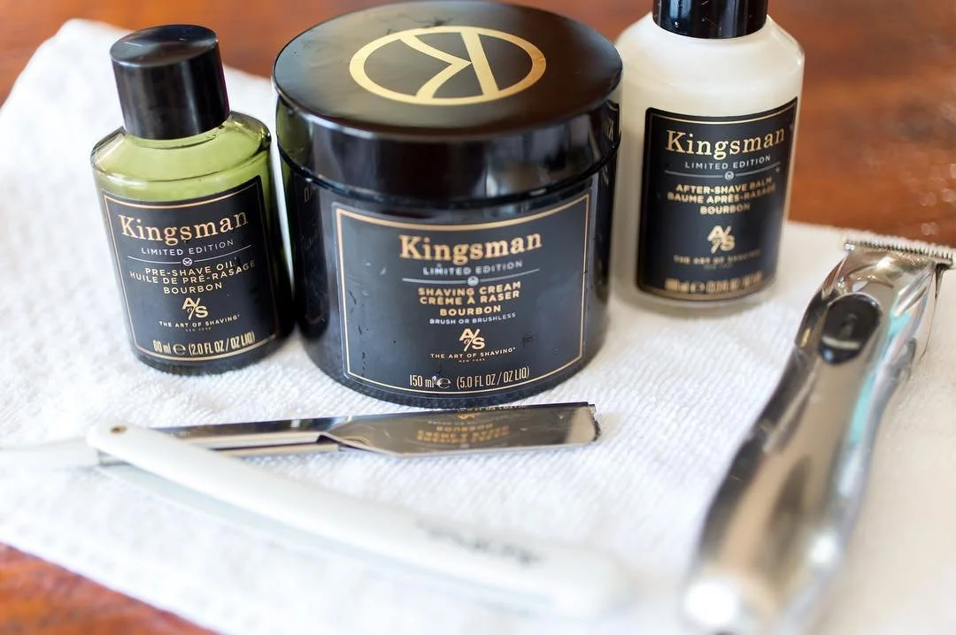 Shaving kit with Kingsman products including pre-shave oil, shaving cream, after-shave balm, straight razor, and electric trimmer on a white towel.
