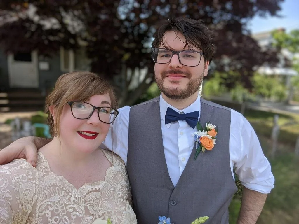 A couple dressed in formal attire, with the woman wearing a lace dress and the man wearing a gray vest and bow tie, poses outdoors with greenery in the background.