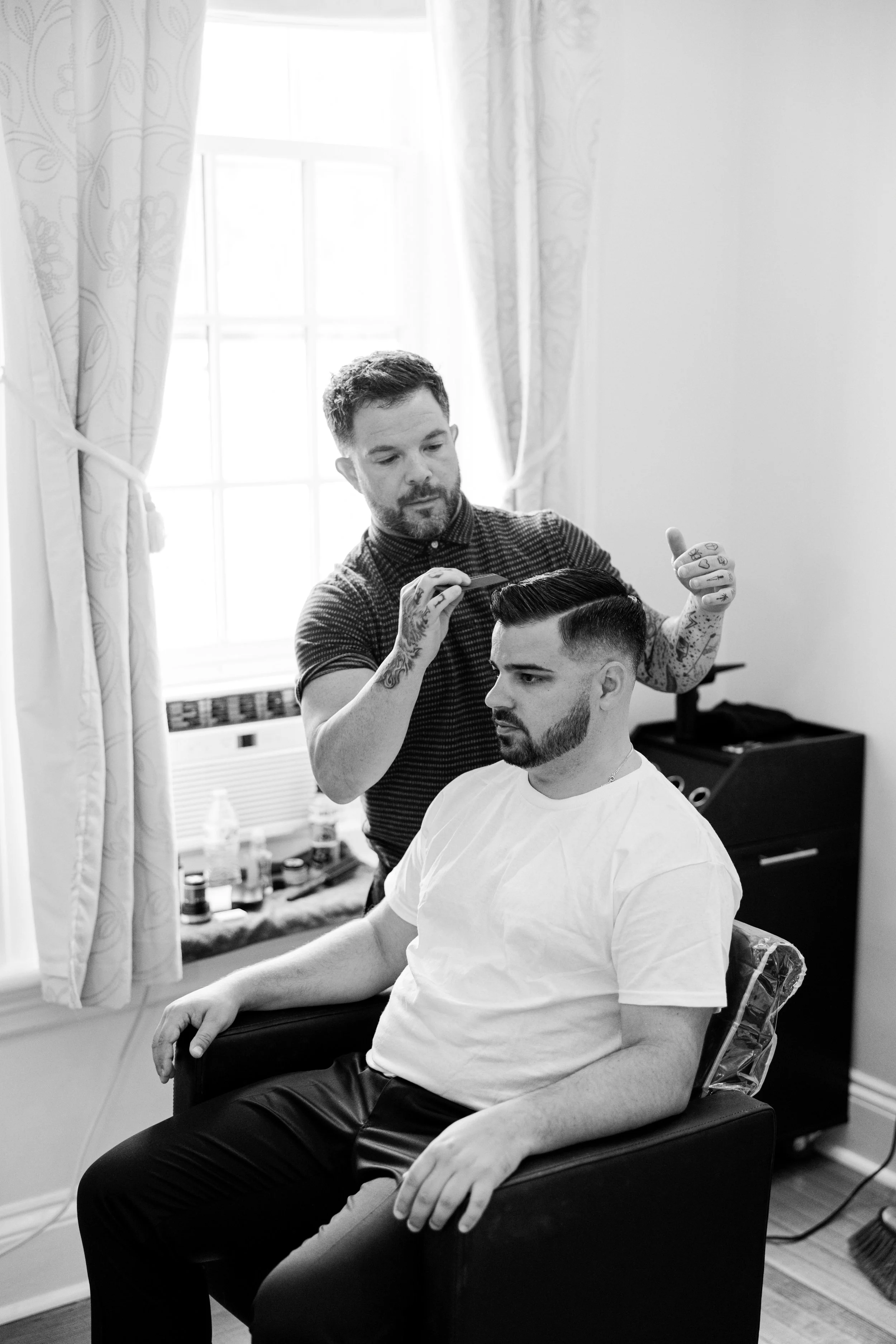 A barber styling a man's hair in a salon chair, both in a room with a large window in the background.