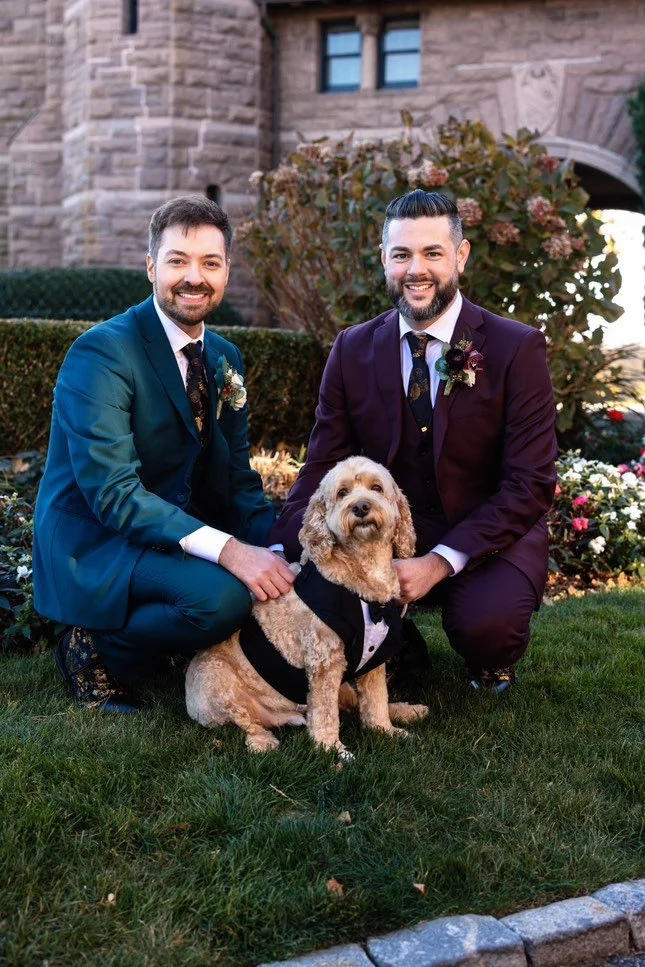 Two men in formal suits pose with a dog wearing a tuxedo outdoors in front of a stone building.
