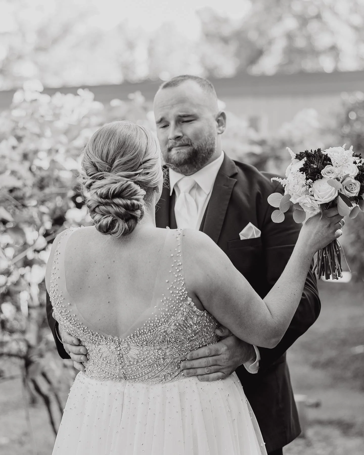 Wedding couple embracing, bride holding bouquet, groom wearing suit, black and white photo.