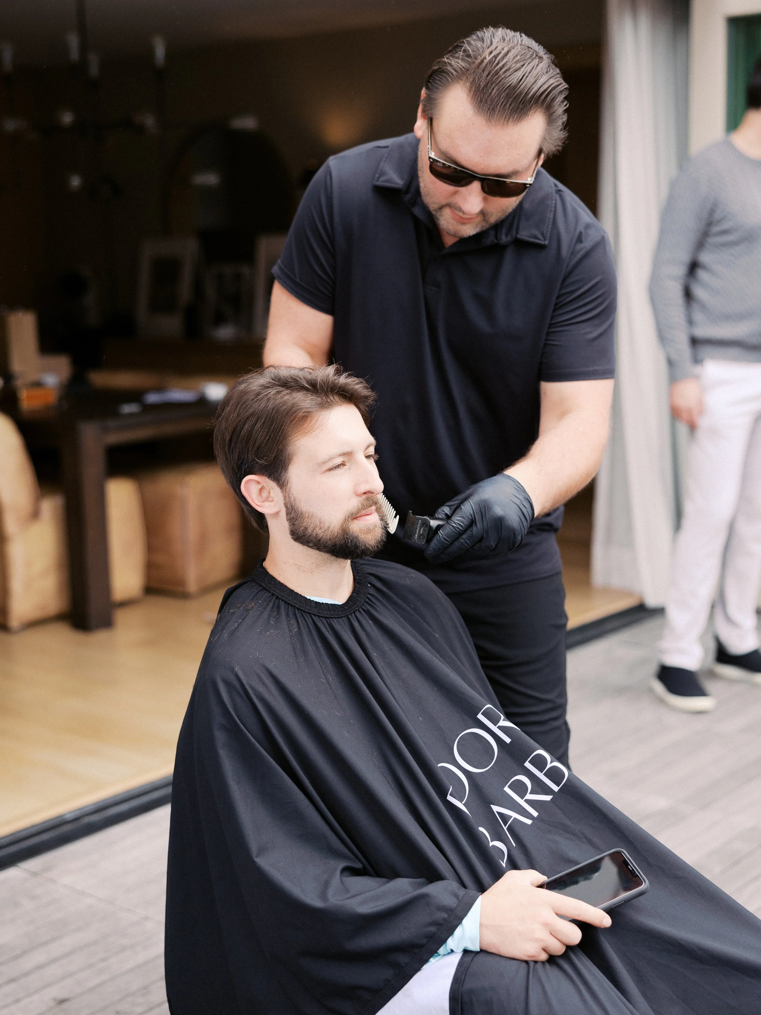 A barber trims a bearded man's facial hair outdoors. The client wears a black barber cape with a phone in his hand. The barber is in a black shirt with gloves.