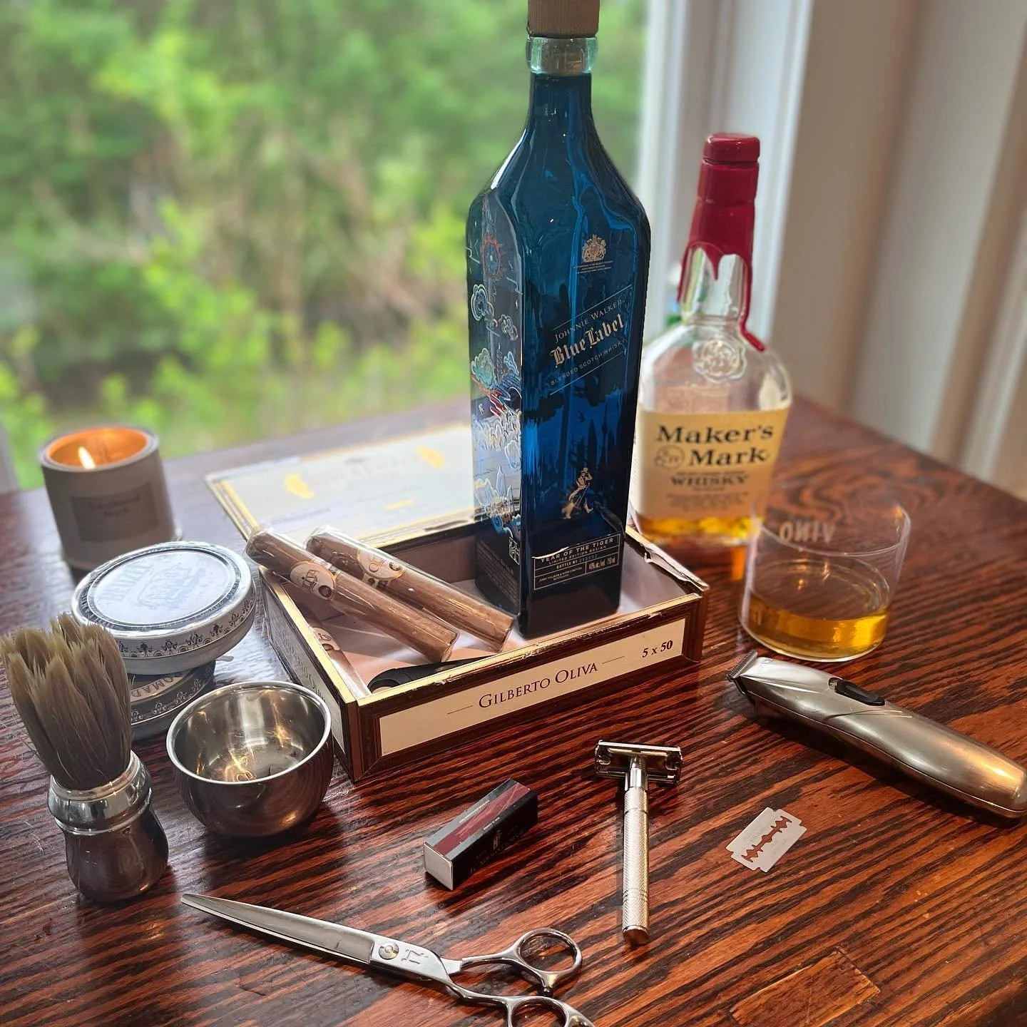 A wooden table with assorted men's grooming items and spirits, including a shaving brush, razor, scissors, a bottle of Johnnie Walker Blue Label, a box with cigars, a candle, and a glass of whiskey.