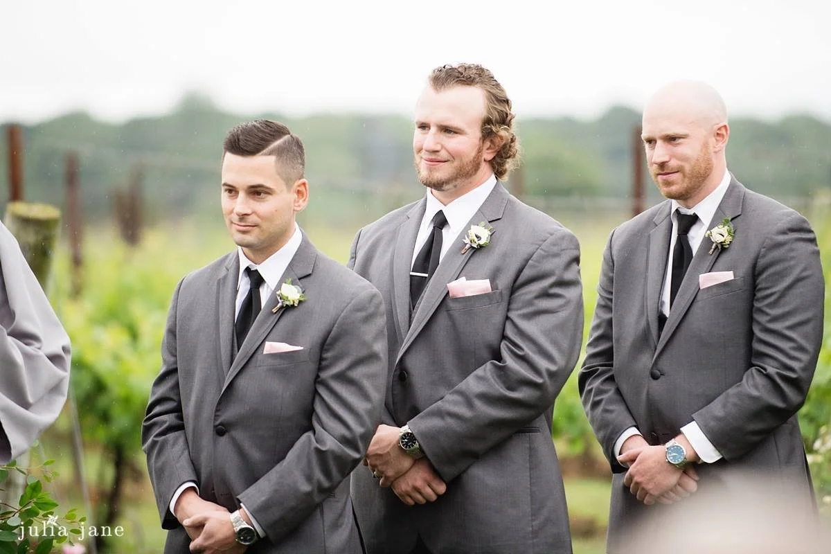 Three groomsmen in gray suits with boutonnieres standing outdoors at a wedding ceremony.