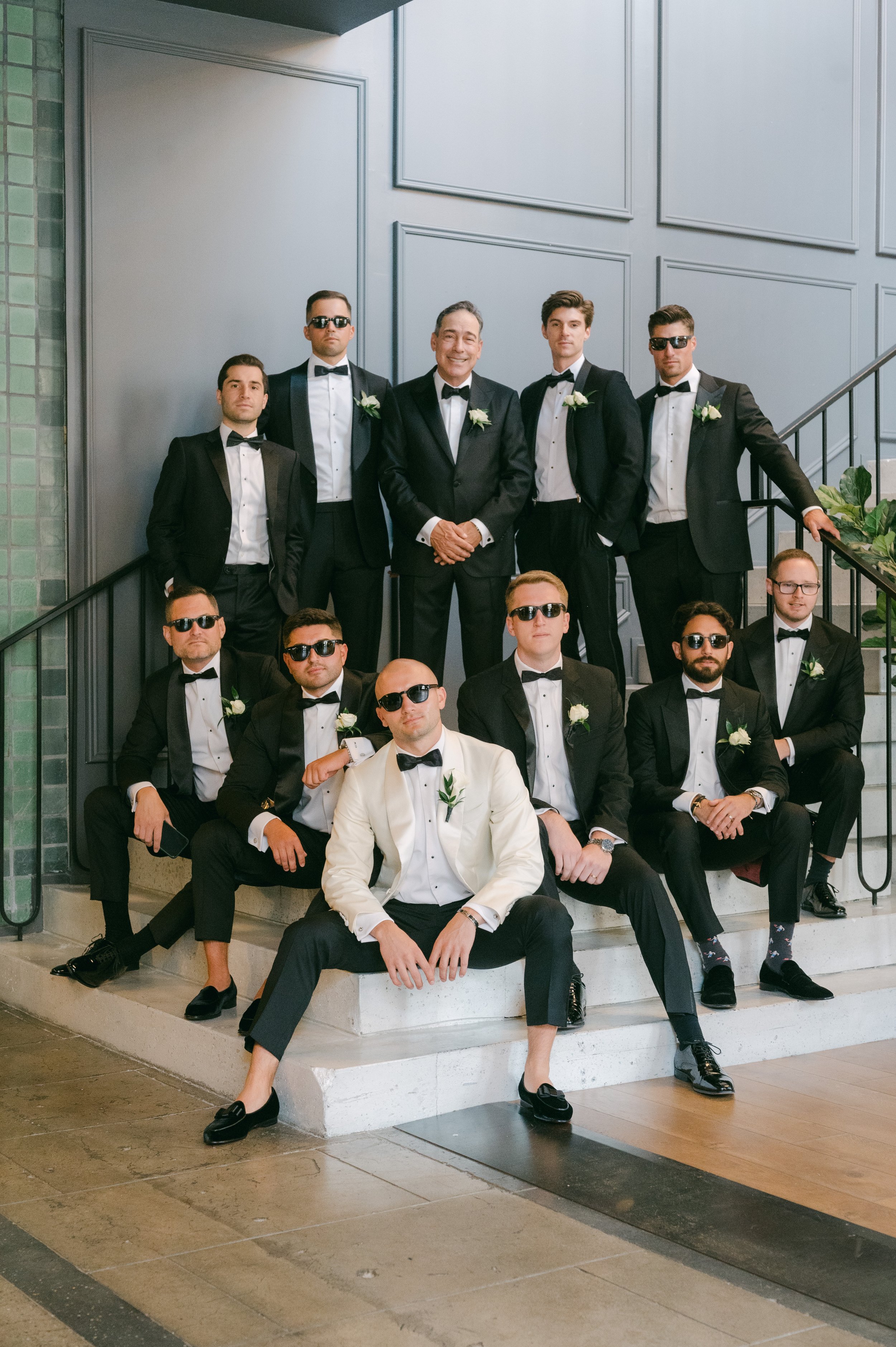 A group of men in formal tuxedos and sunglasses posing on stairs, with one man in a white tuxedo jacket, at a wedding or formal event indoors.