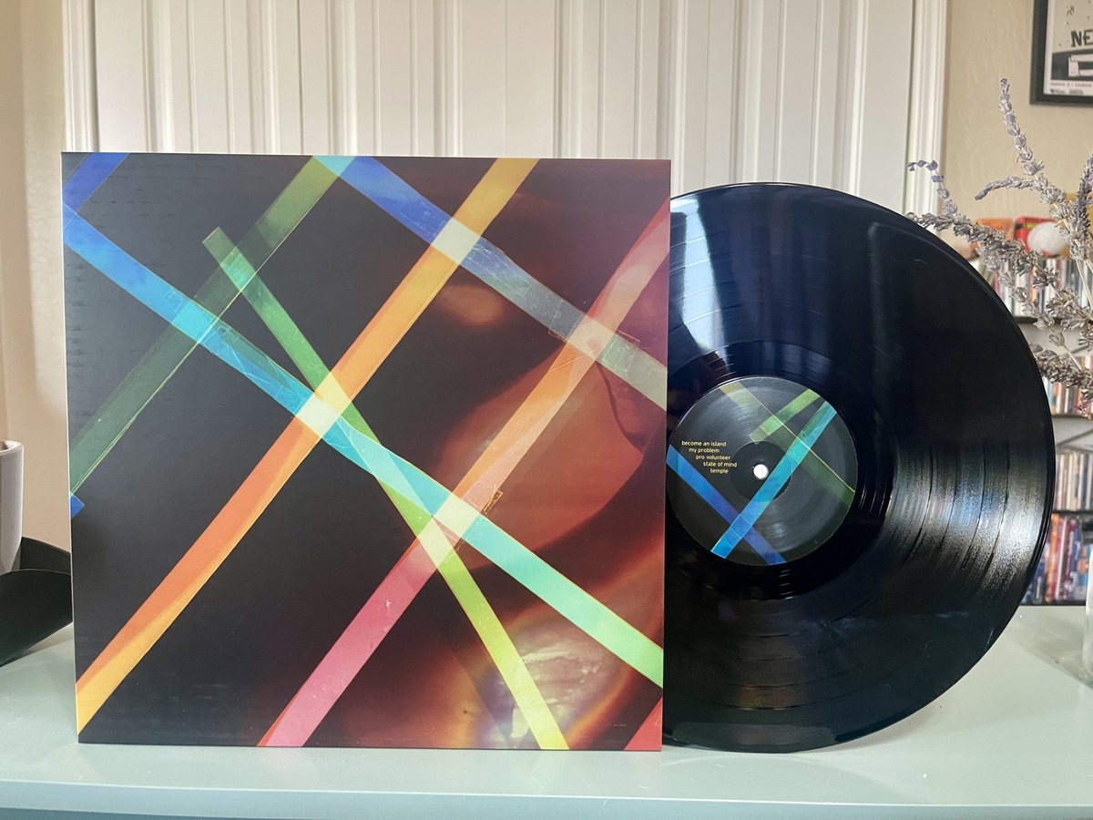 Vinyl record album cover with colorful intersecting lines in green, blue, yellow, and pink on a dark background, placed next to a black vinyl record with a similar colorful label, on a light-colored surface in a room with a patterned wall and a decorated Christmas tree in the background.