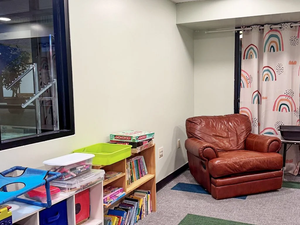 A cozy corner of a children's playroom with a large brown leather armchair beside a window and rainbow-patterned curtains. A small bookshelf filled with children's books and board games sits agains the wall.