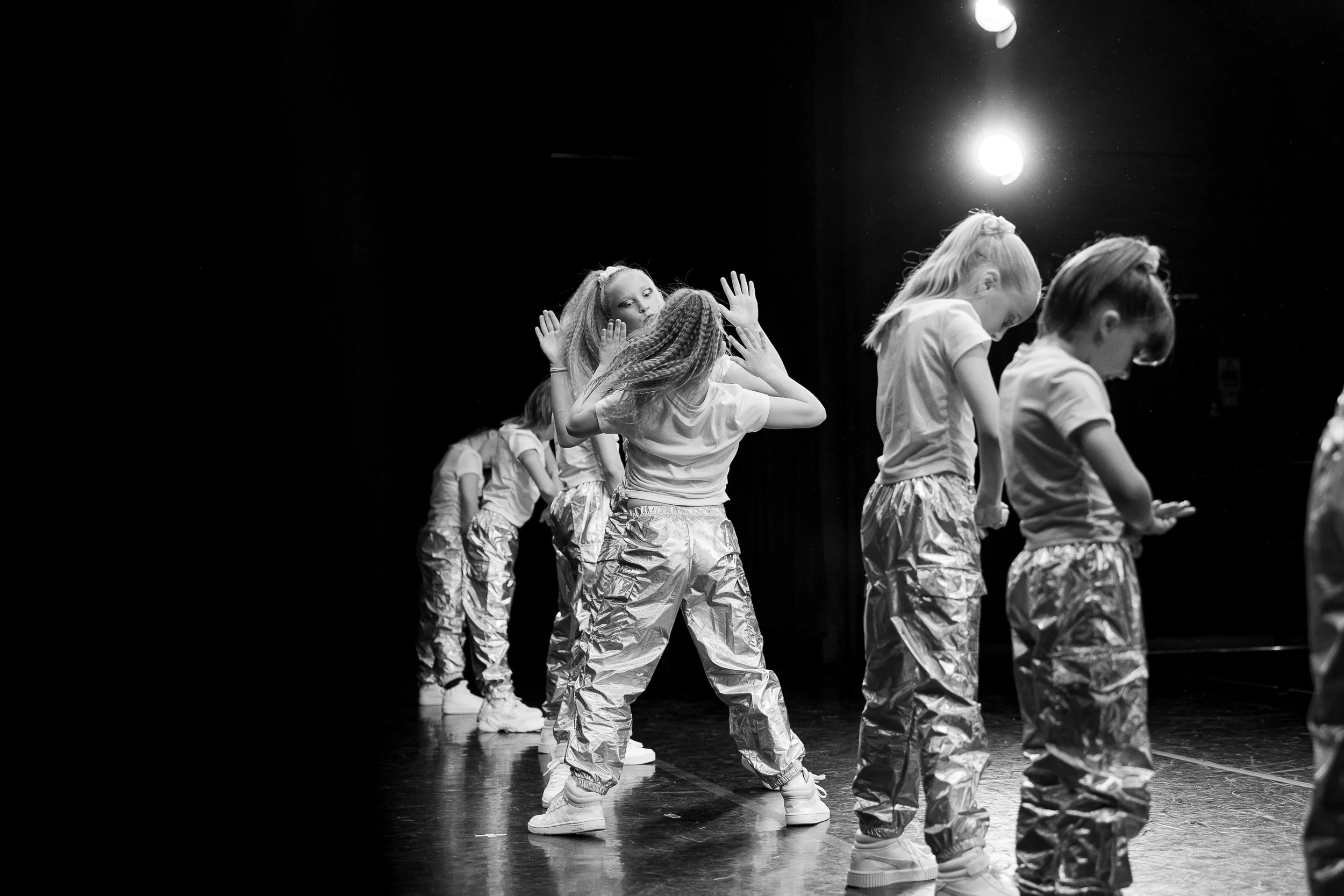 Group of young girls in matching shiny pants and sneakers, participating in a dance or performance on stage with spotlight overhead.