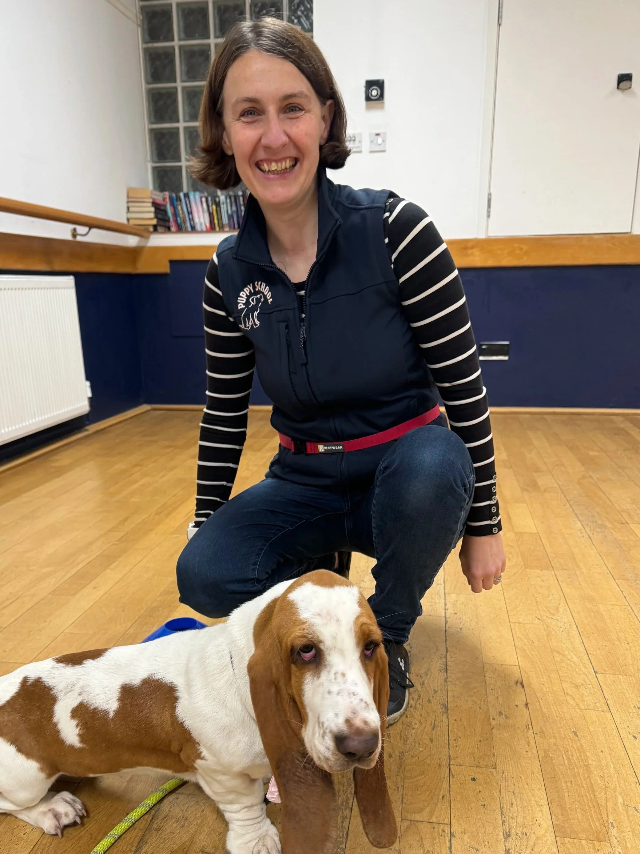 A smiling woman kneeling next to a Basset Hound puppy indoors on a wooden floor, with a window, books, and radiators in the background.