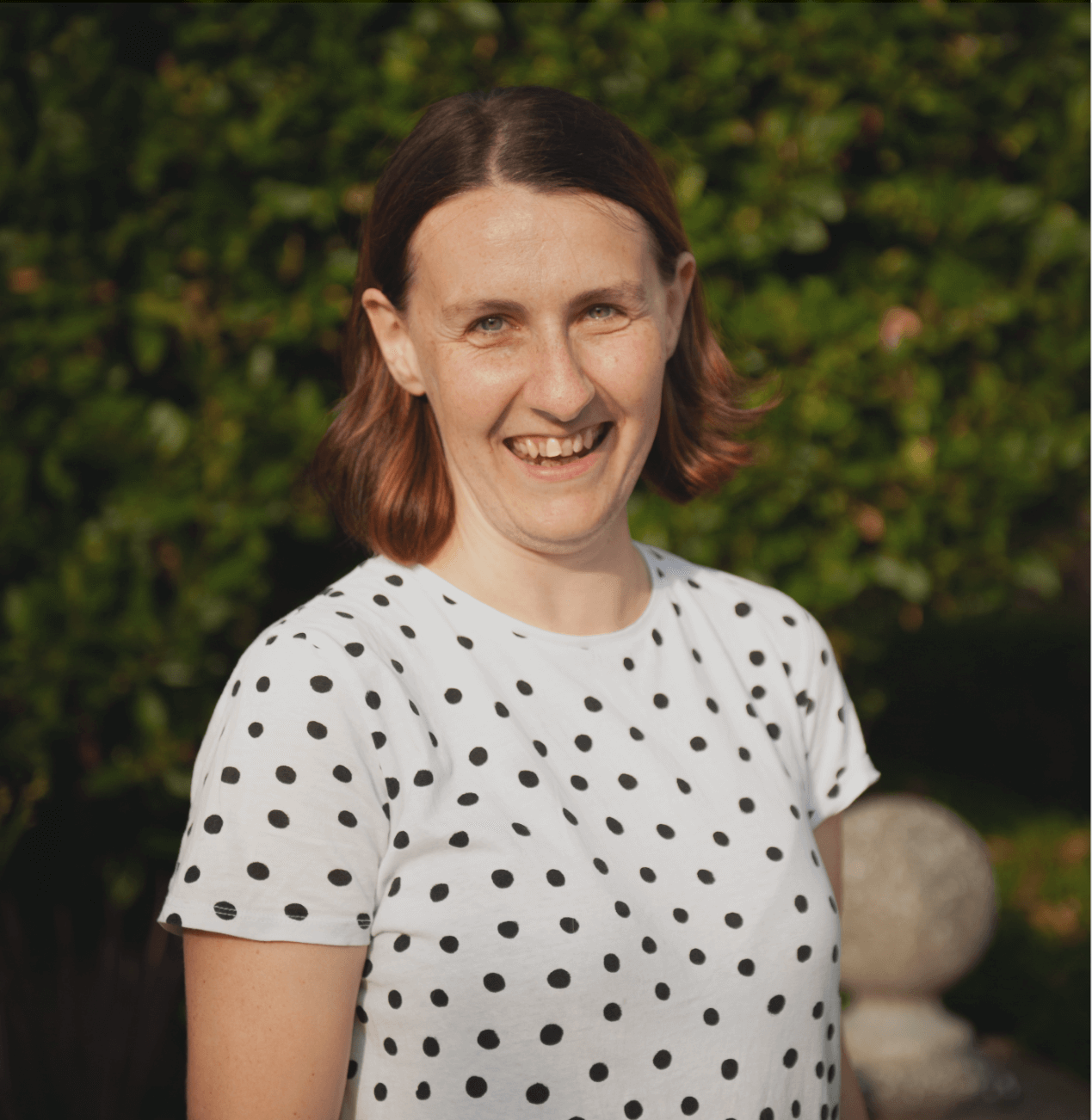 A woman with shoulder-length brown and reddish hair smiling and wearing a white t-shirt with black polka dots, standing outdoors in front of green foliage.