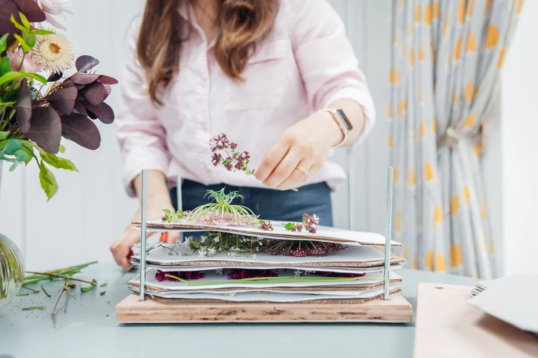 Lucy placing fresh flowers into a flower press. She is blurred in the background whilst the press is in focus.