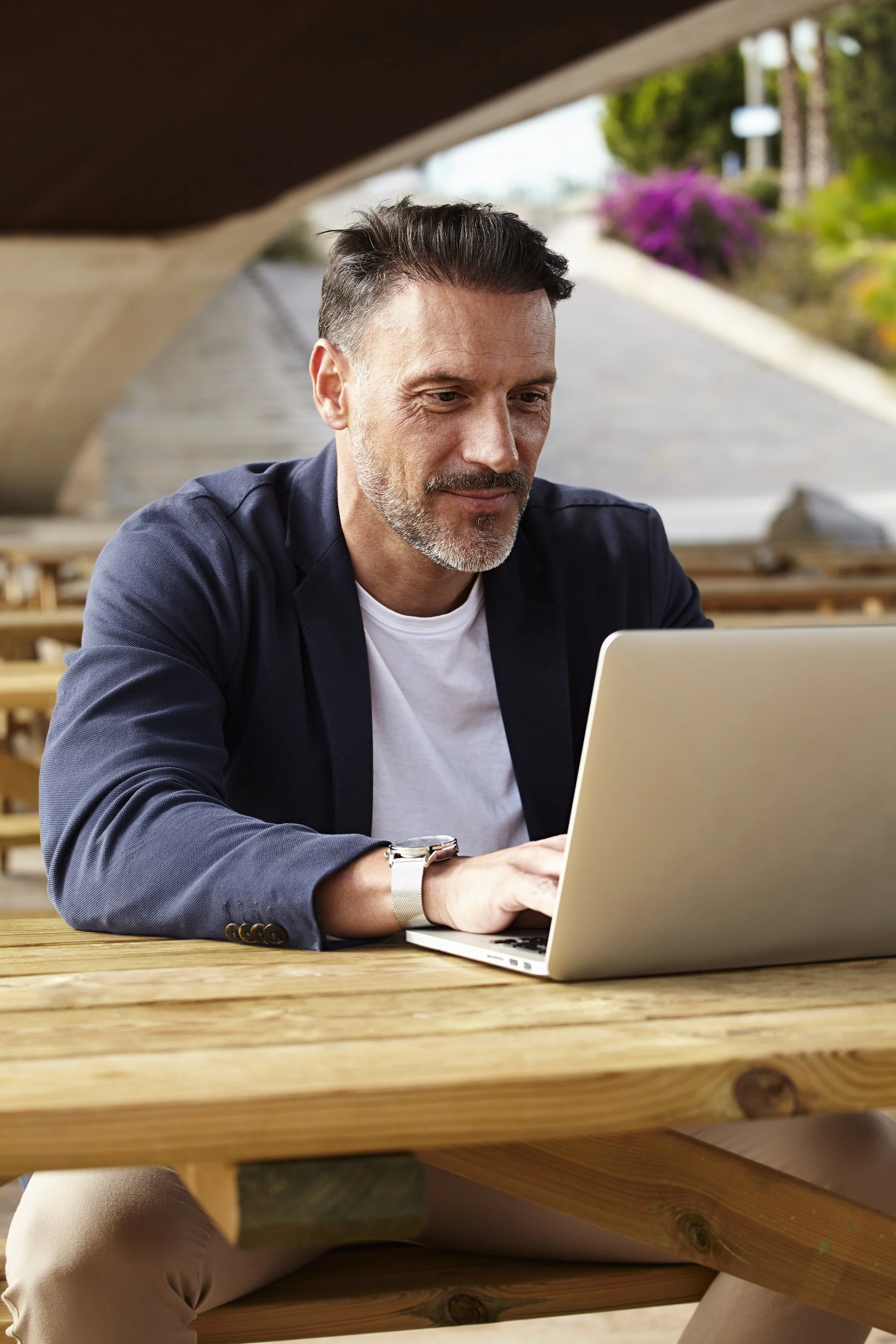 A man with dark hair and a beard sitting at a wooden outdoor table using a silver laptop on a 1 on 1 retirement coaching call
