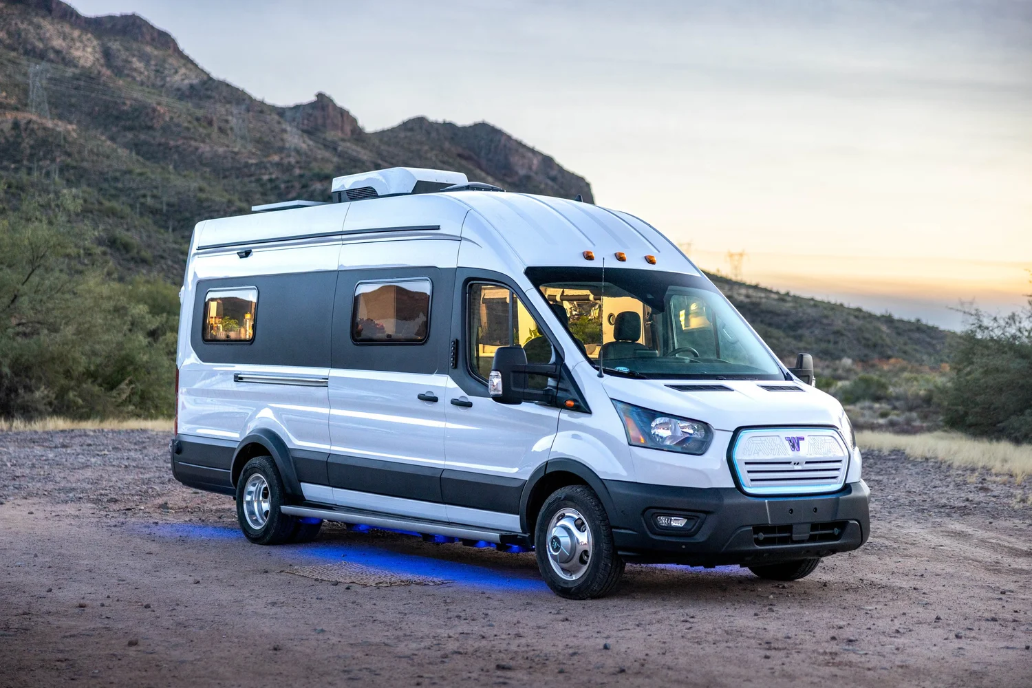 A white camper van parked on a dirt area with desert hills in the background, during sunset.
