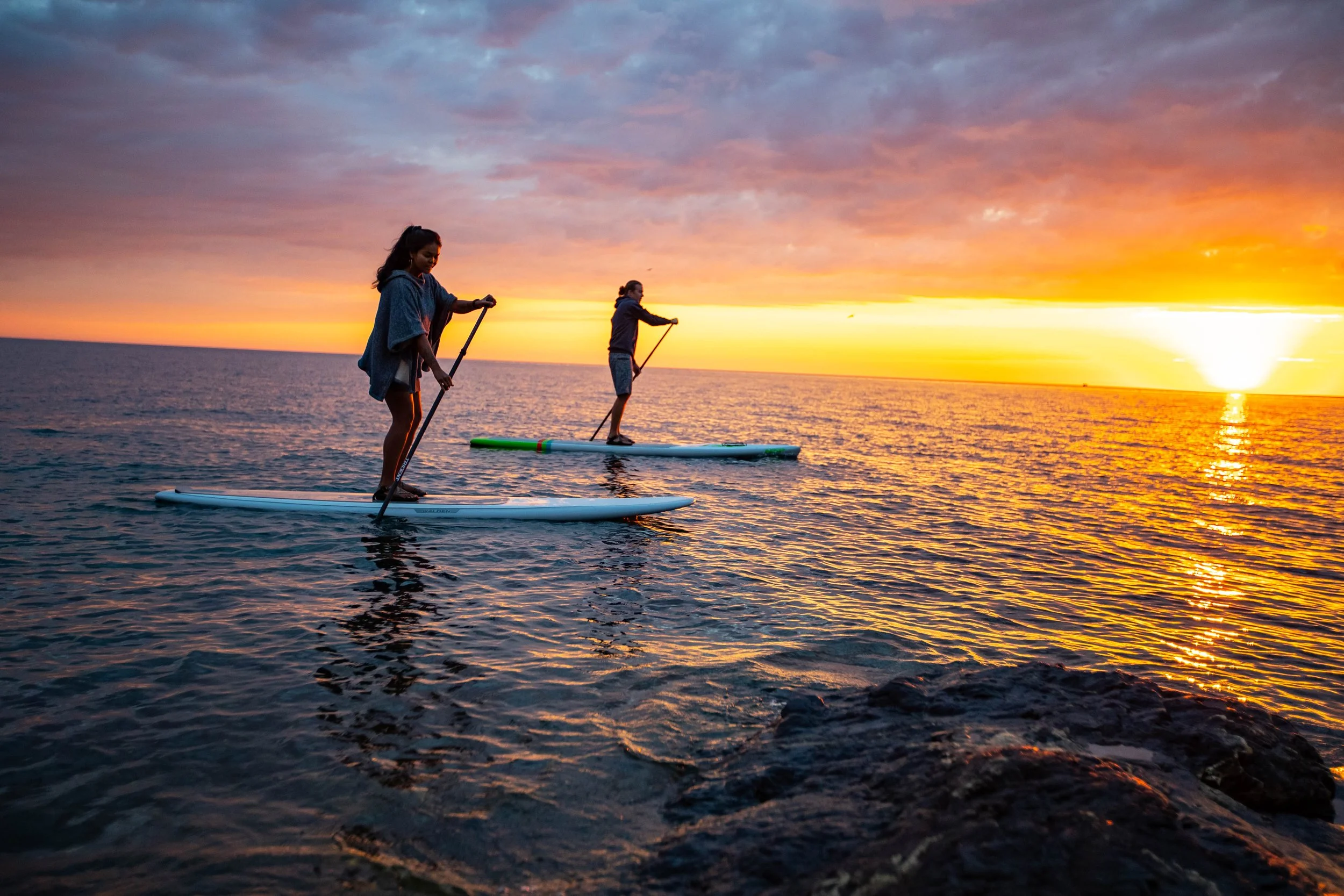 Two people stand on paddleboards in the water during sunset, with colorful clouds and the sun setting on the horizon.