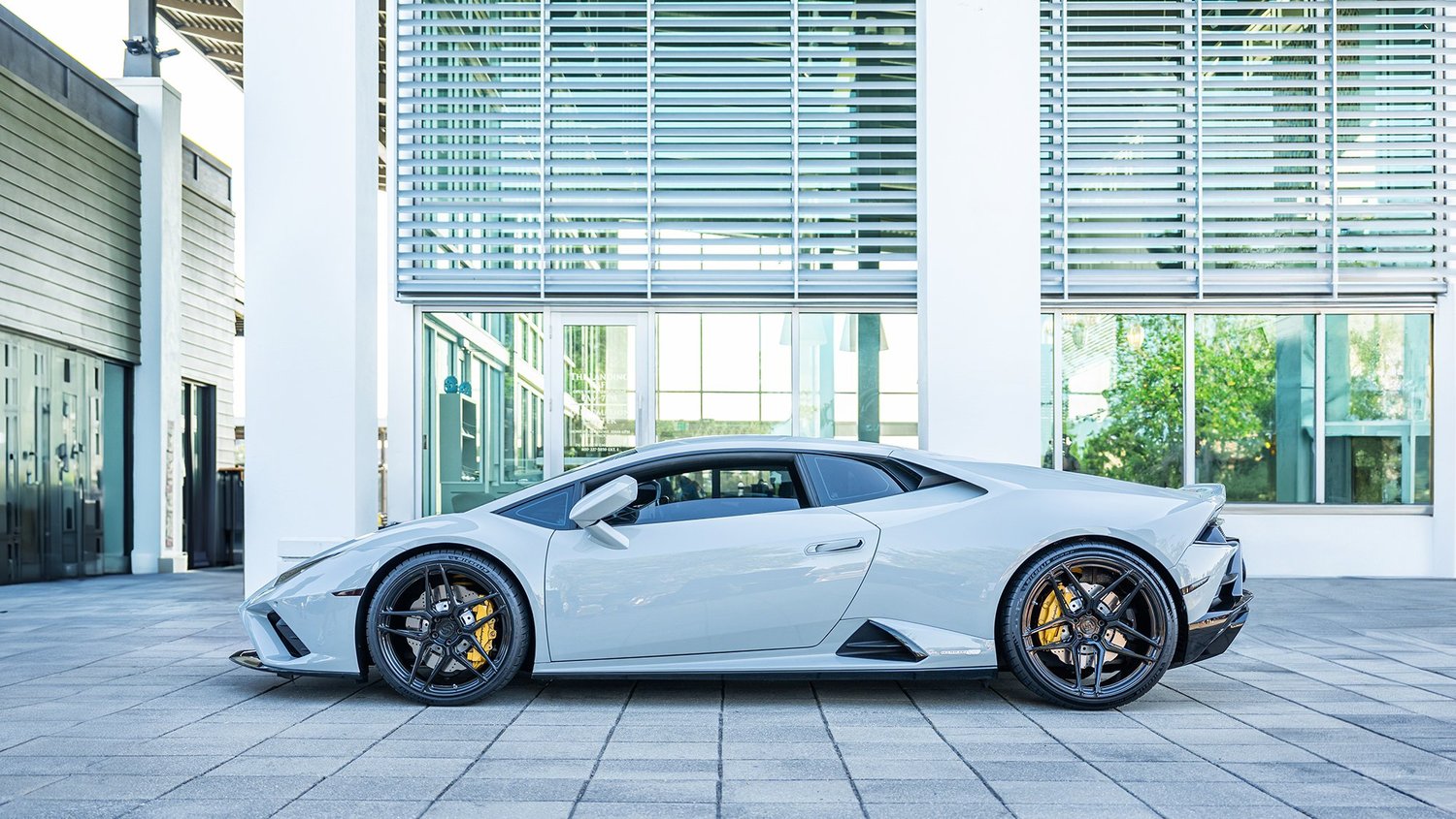 A silver Lamborghini sports car parked in front of a modern building with large glass windows and white and green exterior.