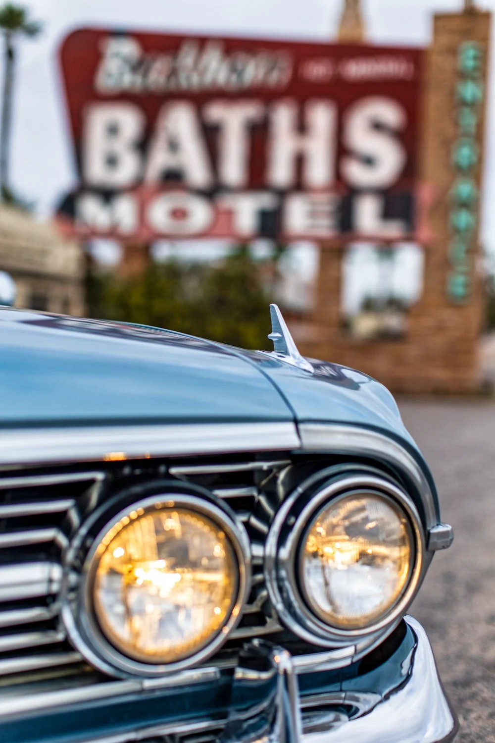 Close-up of a vintage car with illuminated headlights parked in front of a blurred sign that reads "BATHS MOTEL" in the background.