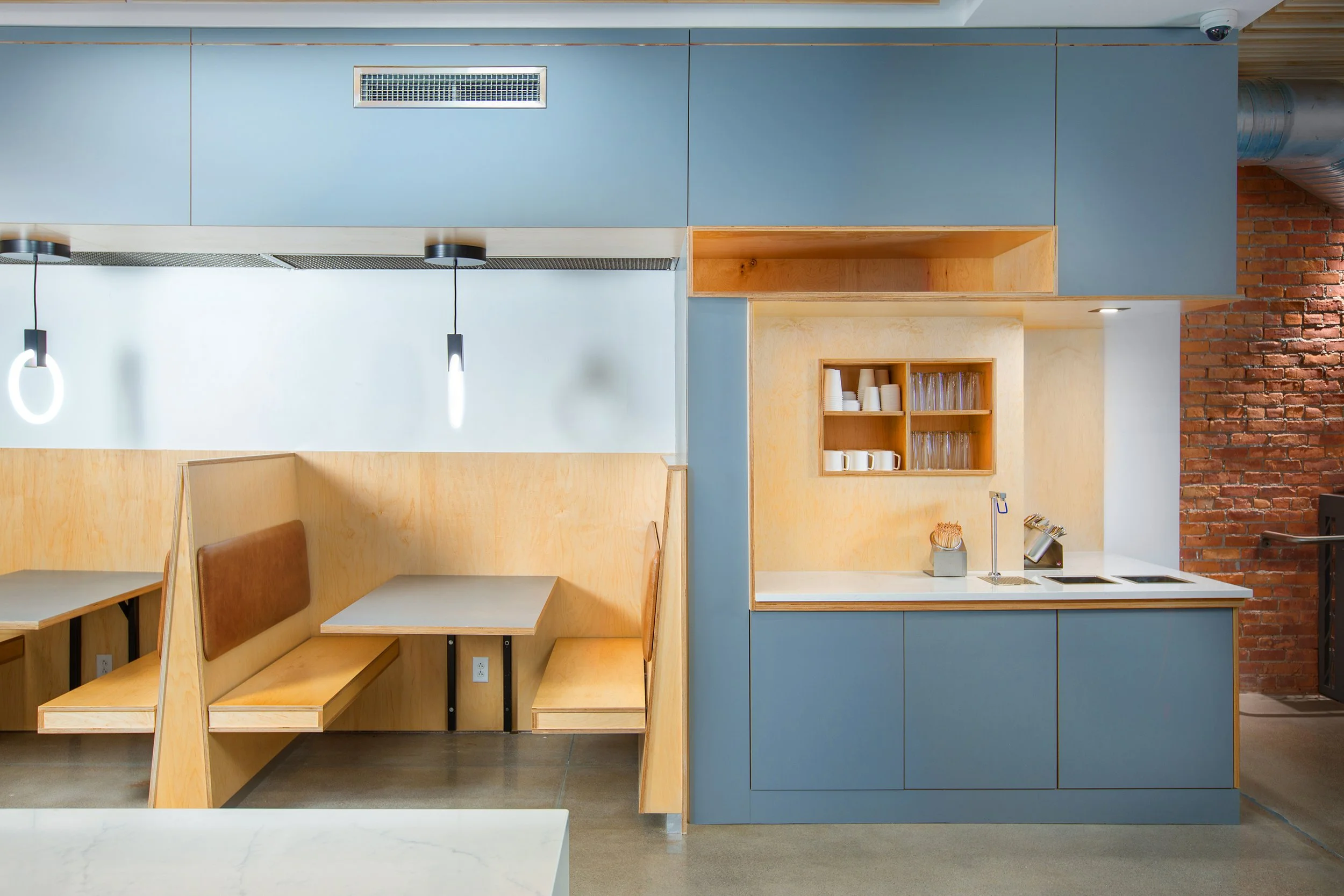 Modern cafe seating area with booths on the left featuring wood paneling and upholstered cushions. On the right, a kitchen counter with a blue base, open shelves with cups and glasses, a sink, and a brick wall in the background. Pendant lights above 