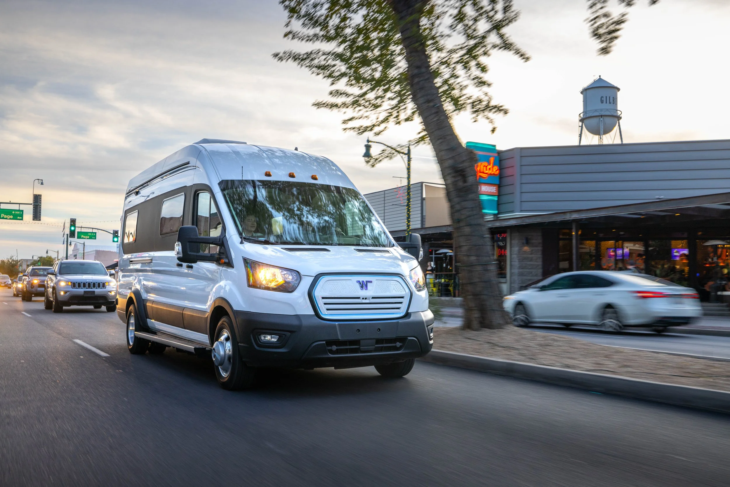 A white van driving on a city street during dusk with buildings, restaurants, and cars visible in the background.